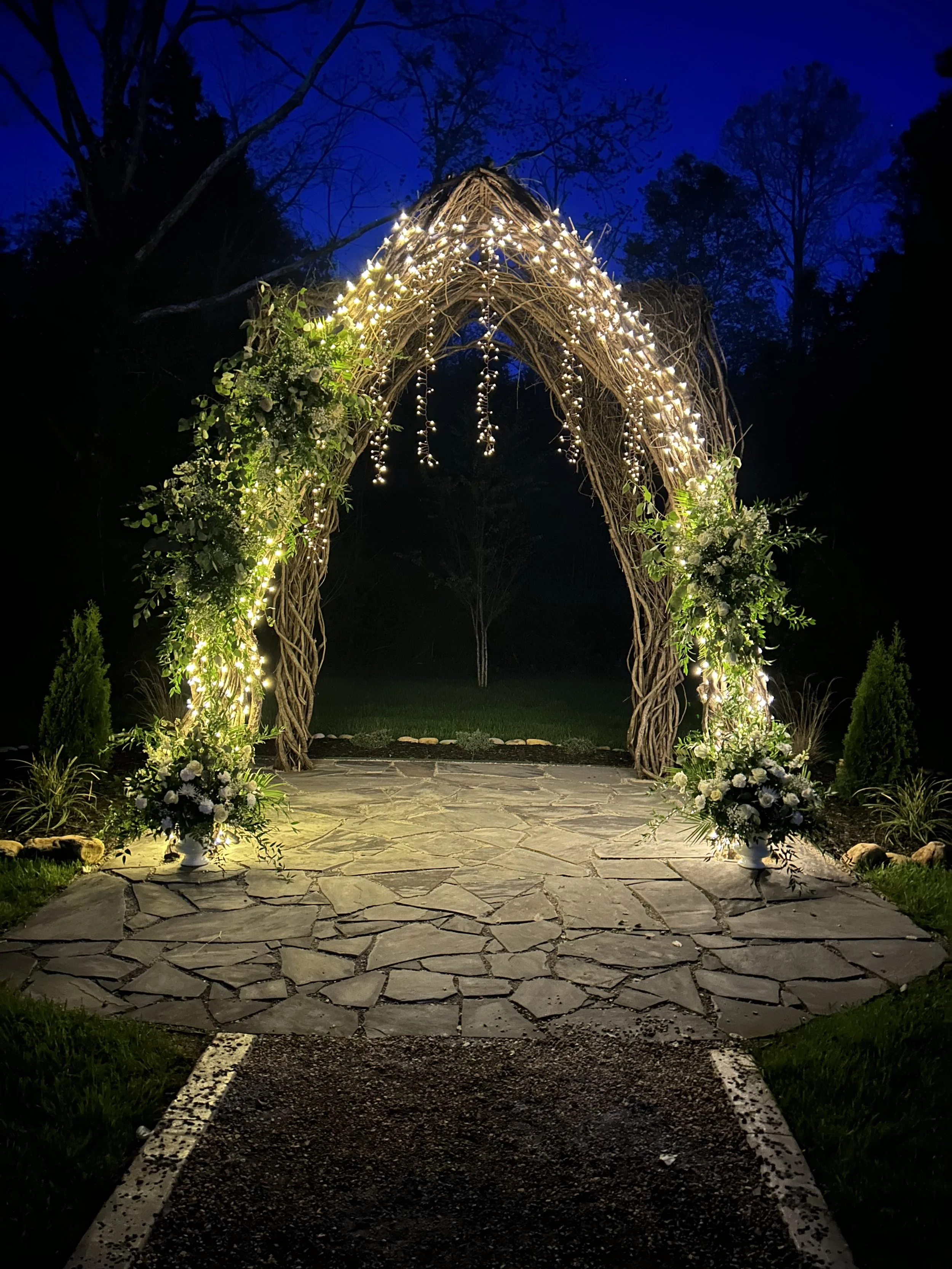 A decorative outdoor wedding arch in East Tennessee made of intertwined branches, illuminated with string lights, decorated with white flowers, and set up on a stone pathway at night.