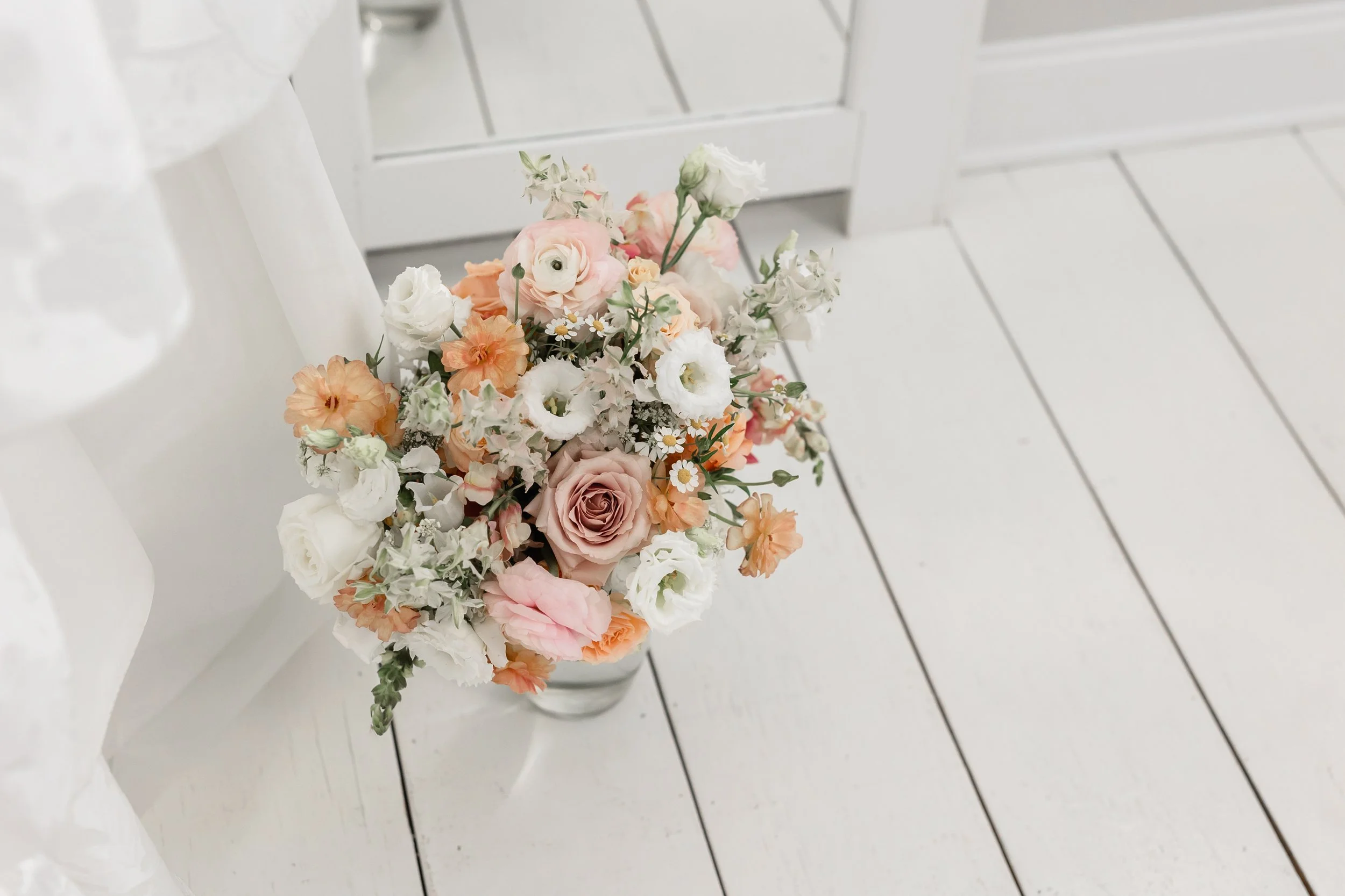 Spring wedding bouquet with peach, blush, and white flowers arranged beside a wedding dress on white wood floors at Ramble Creek.