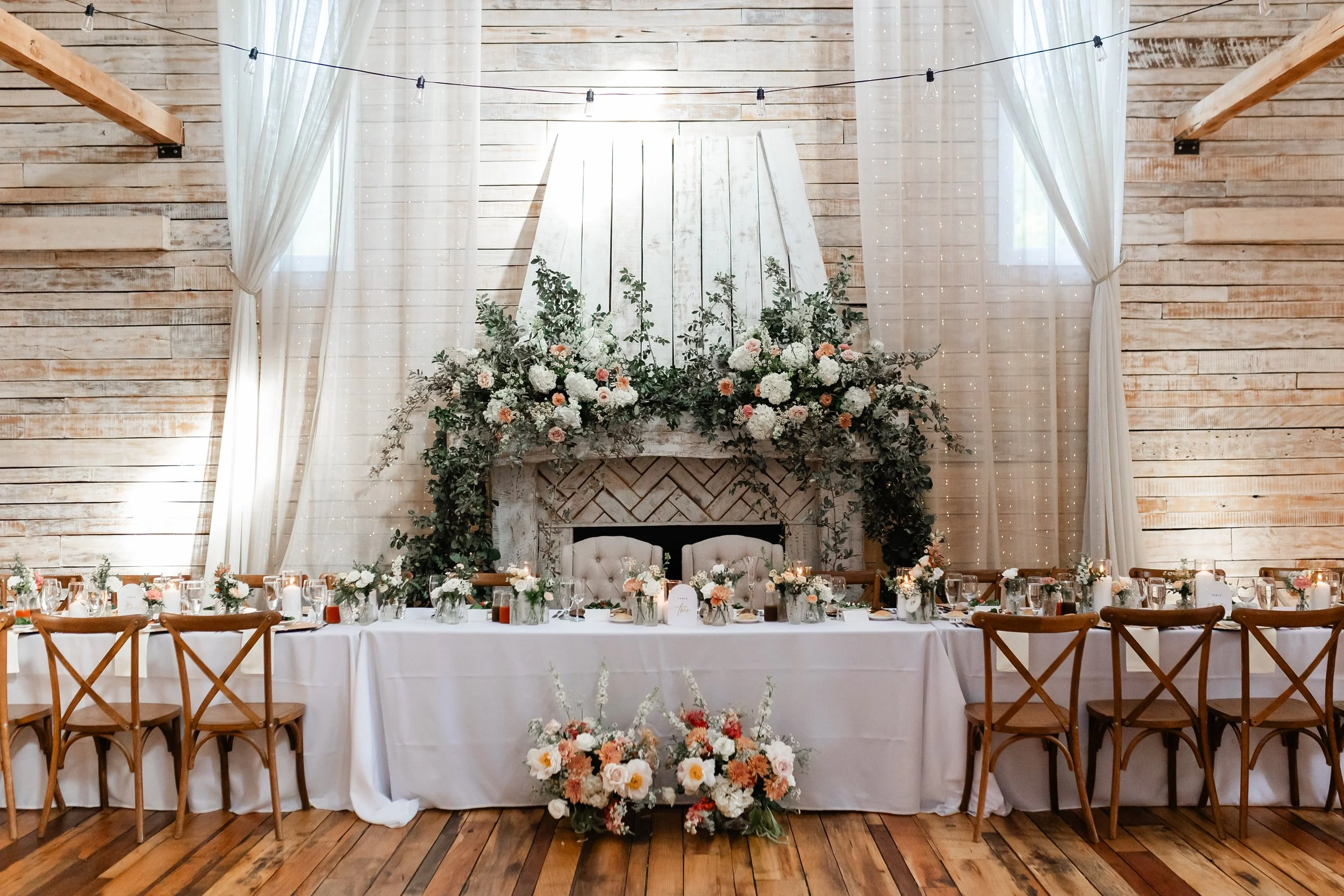 Ramble Creek wedding reception head table inside Ramble Creek’s Grand Hall, featuring white drapery, wood plank walls, cross-back chairs, candles, and lush peach, white, and greenery floral arrangements around the fireplace backdrop.