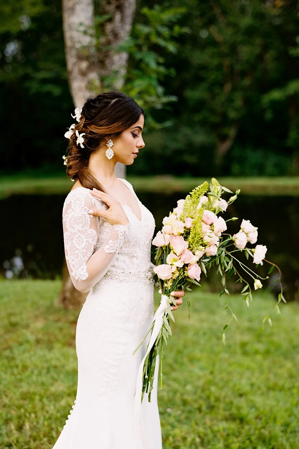 A bride in a white lace wedding dress holding a bouquet of pink and cream flowers, standing outdoors near a lake surrounded by trees at Ramble Creek, the best all-inclusive private wedding venue in Tennessee.