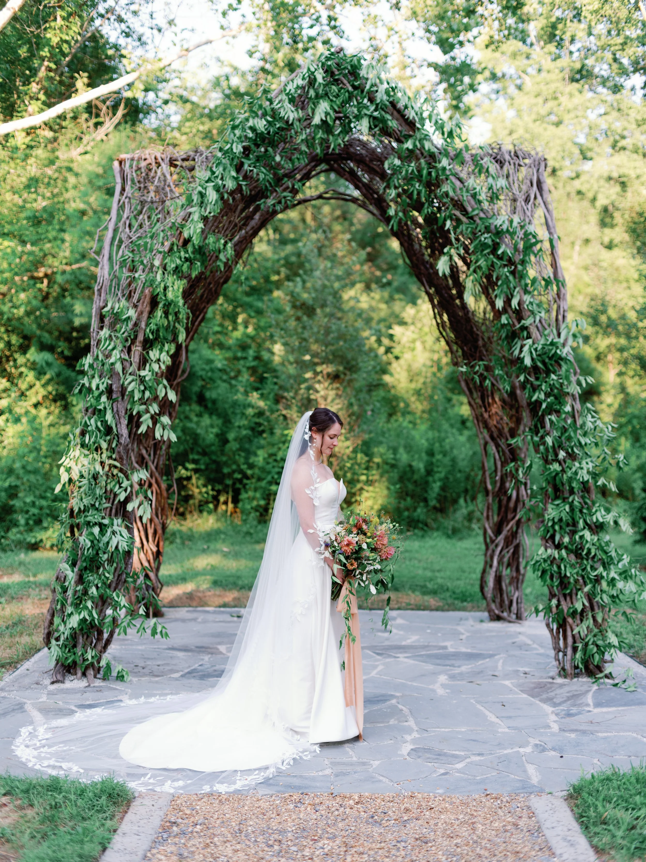 A bride in a white wedding dress holding a bouquet standing under a decorative arch made of intertwined branches and green leaves, in an outdoor setting with trees and greenery in the background.