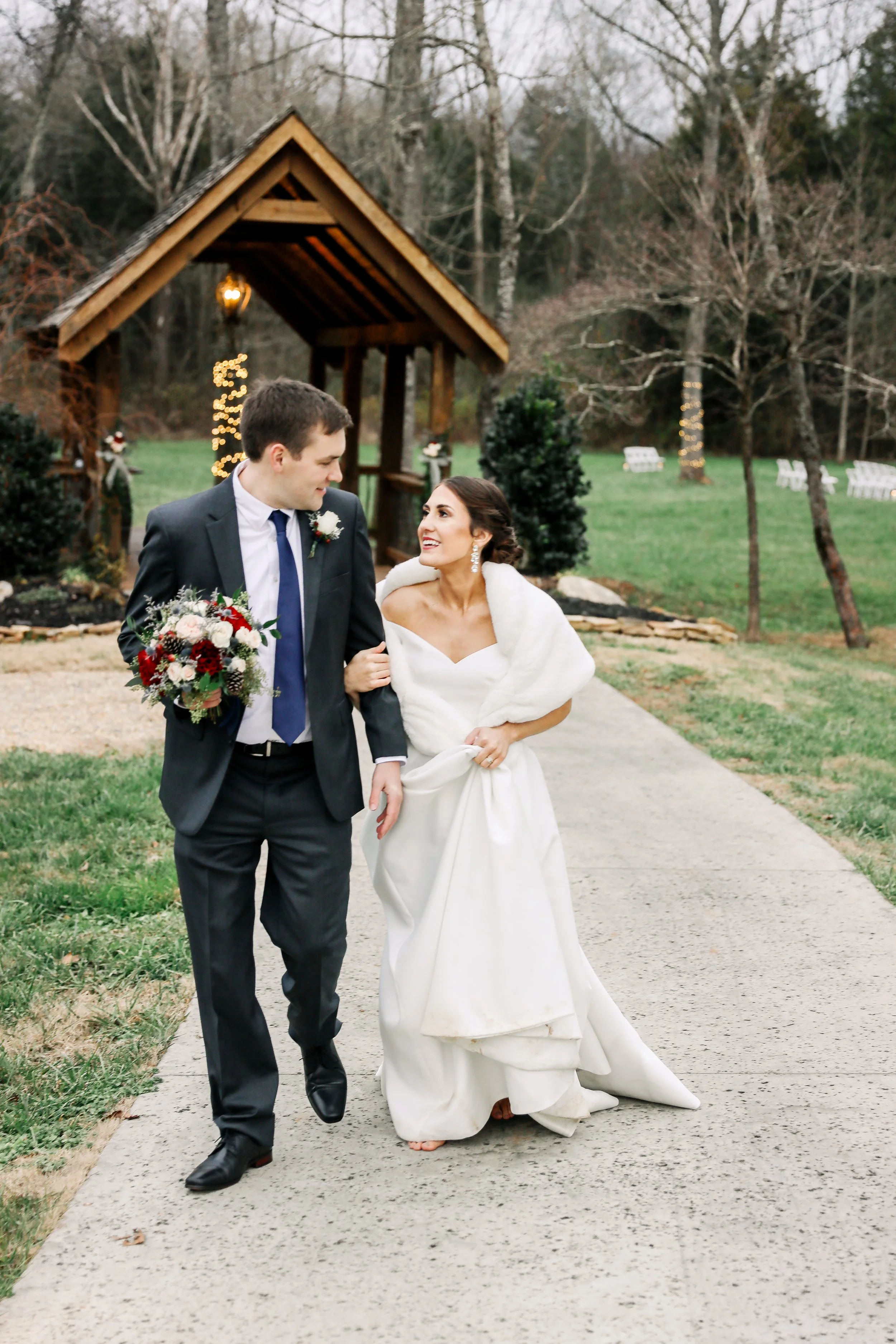 Bride and groom walking together at Ramble Creek in winter, with the bride wearing a white faux fur wrap near the outdoor ceremony pavilion