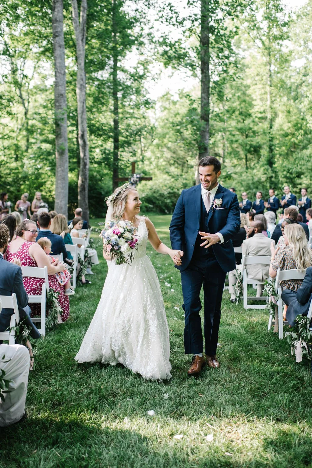 A bride and groom walking hand in hand down an outdoor aisle during a wedding ceremony in a forested area, smiling and happy surrounded by guests seated on both sides at Ramble Creek all-inclusive wedding venue.