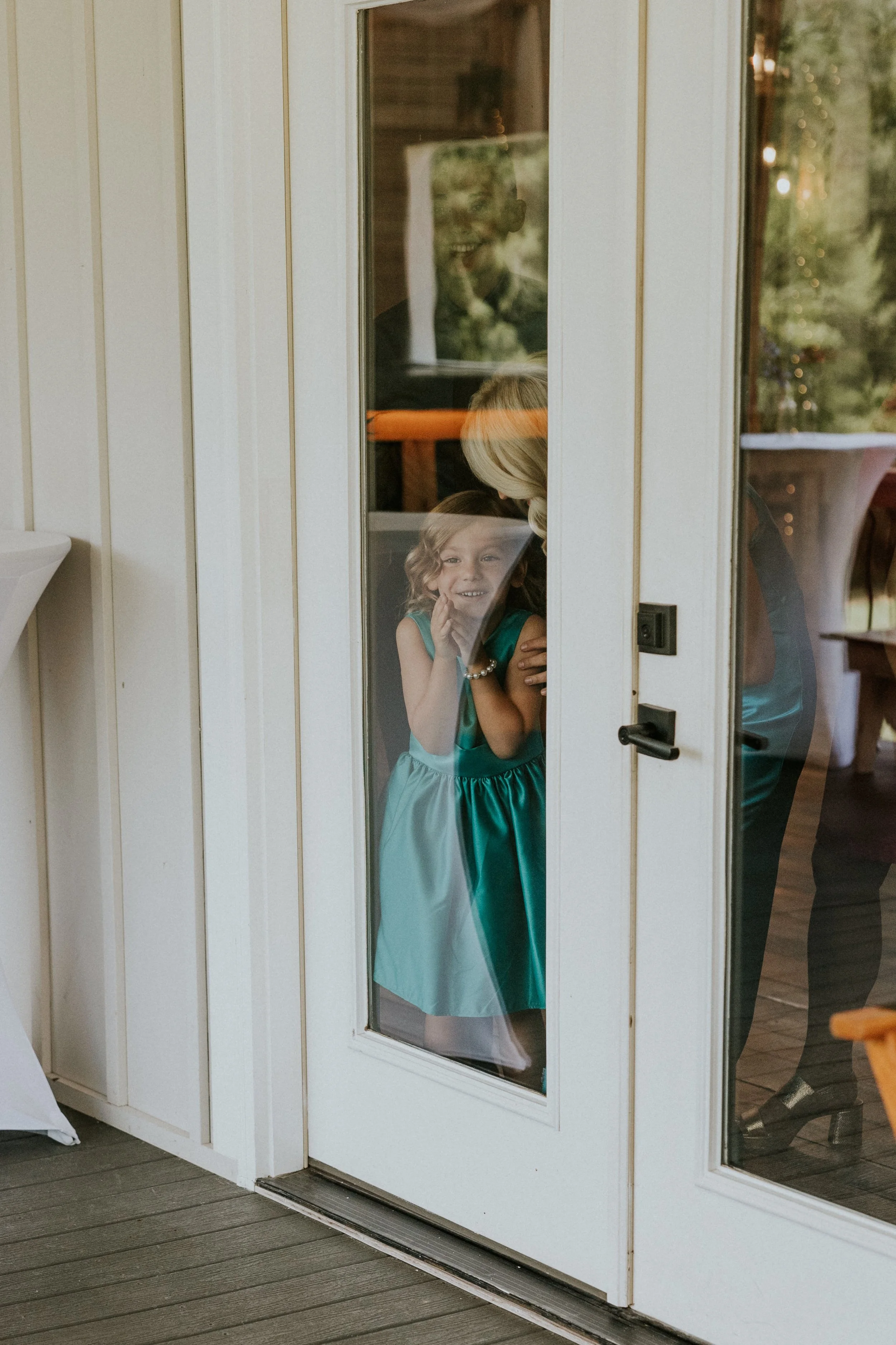 A young flower girl in a teal dress smiling and looking out through a glass door at a wedding or celebration, with a woman beside her and a man in the background.