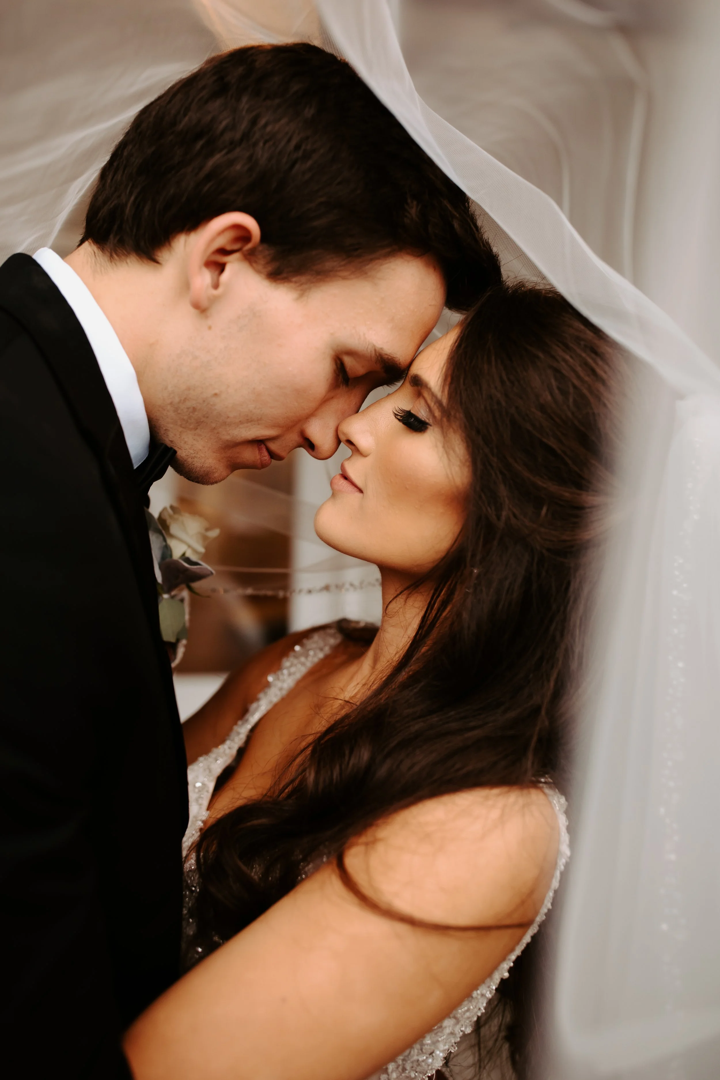 Bride and groom sharing an intimate forehead touch beneath a soft wedding veil, highlighting the bride’s long dark hair and bridal makeup.