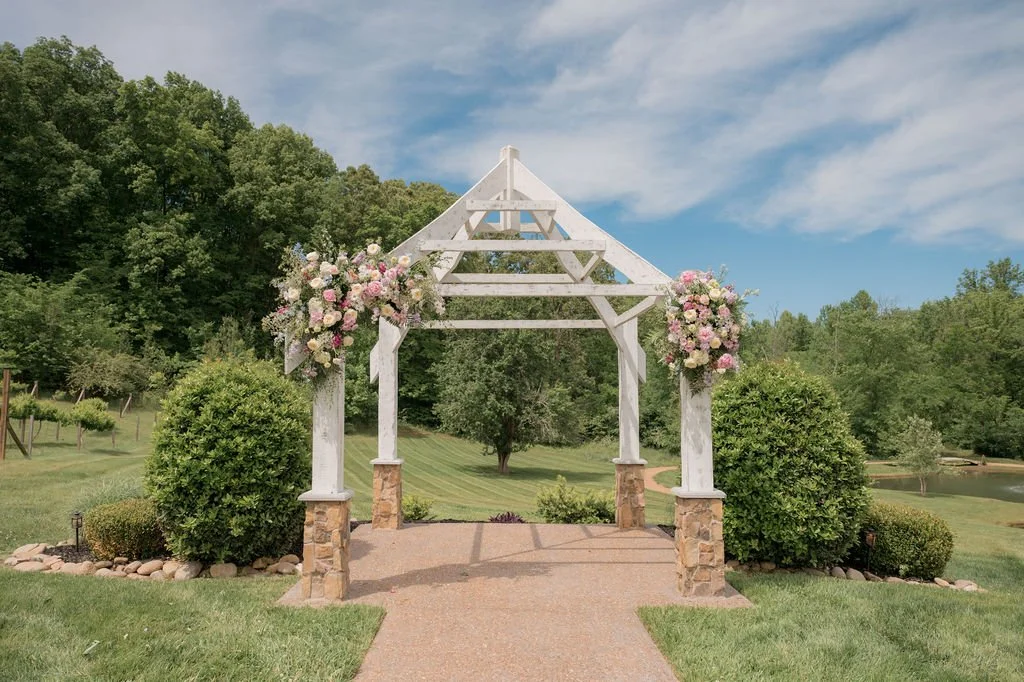 Circular photo of the Upper Lawn ceremony site at Ramble Creek featuring an outdoor wooden arbor decorated with colorful floral arrangements and chandeliers, where a bride and groom stand together beneath the structure surrounded by lush greenery