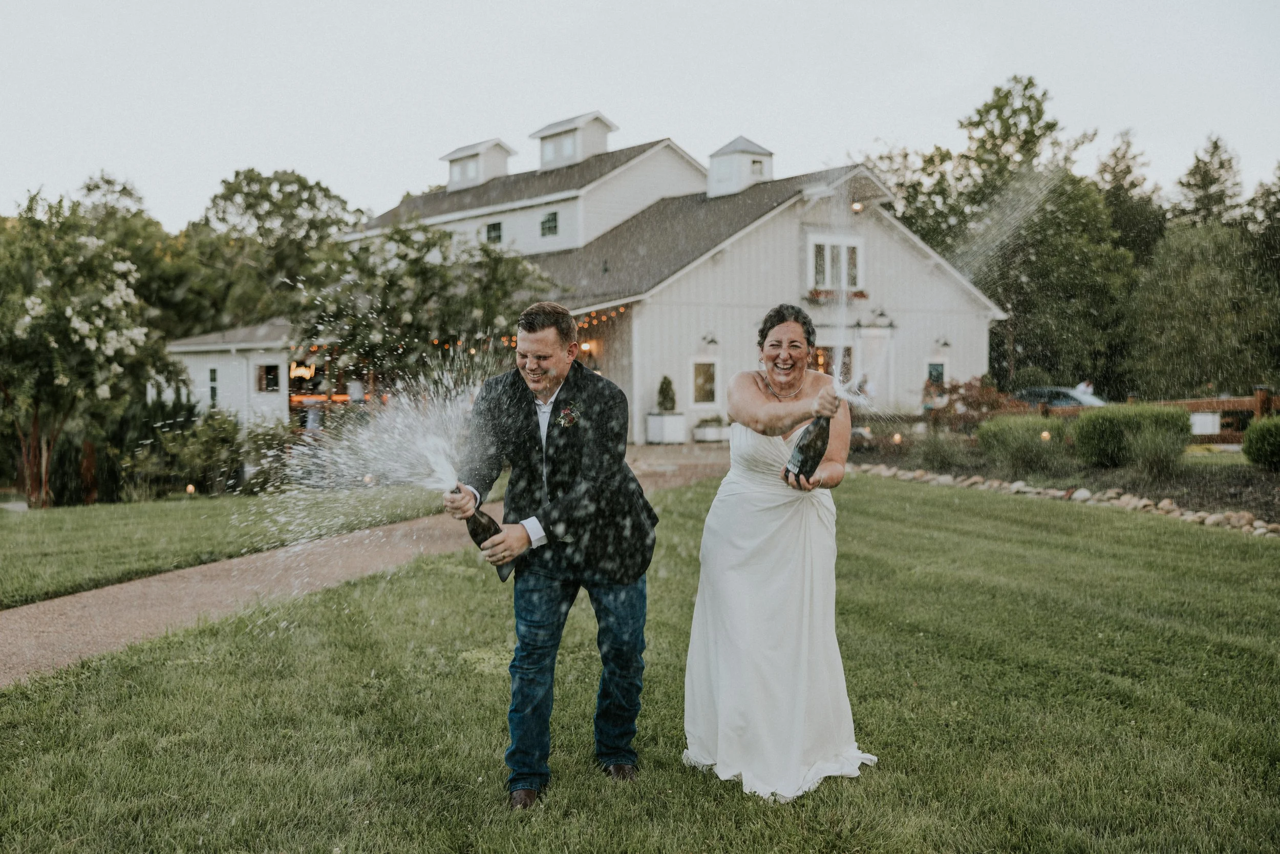 Couple spraying champagne on the upper lawn at Ramble Creek, a Tennessee wedding venue with a white barn, landscaped grounds, and joyful outdoor wedding scenery
