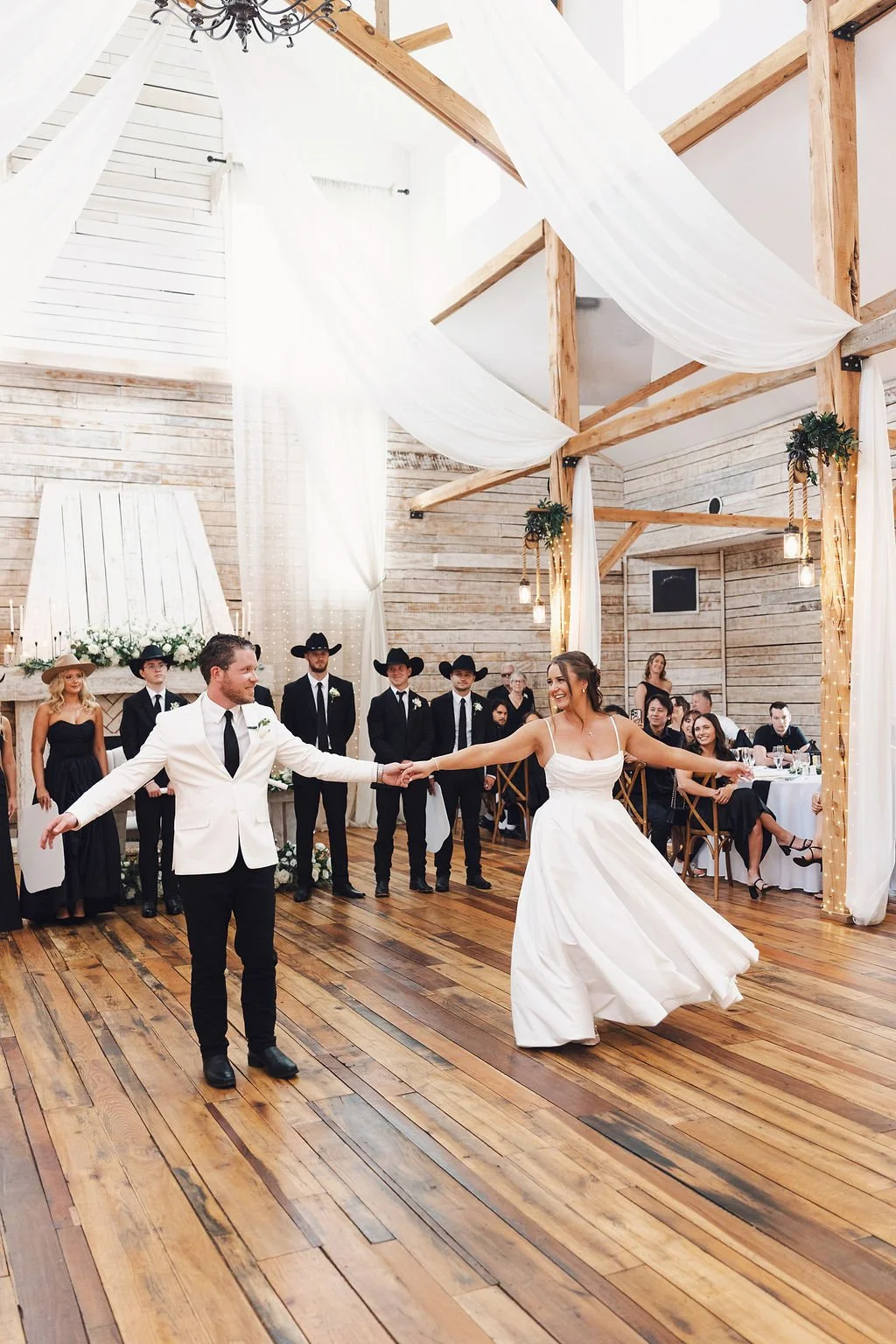 Bride and groom share their first dance inside a bright rustic wedding reception venue with white ceiling drapes, wood beams, chandeliers, and guests watching from the dance floor.