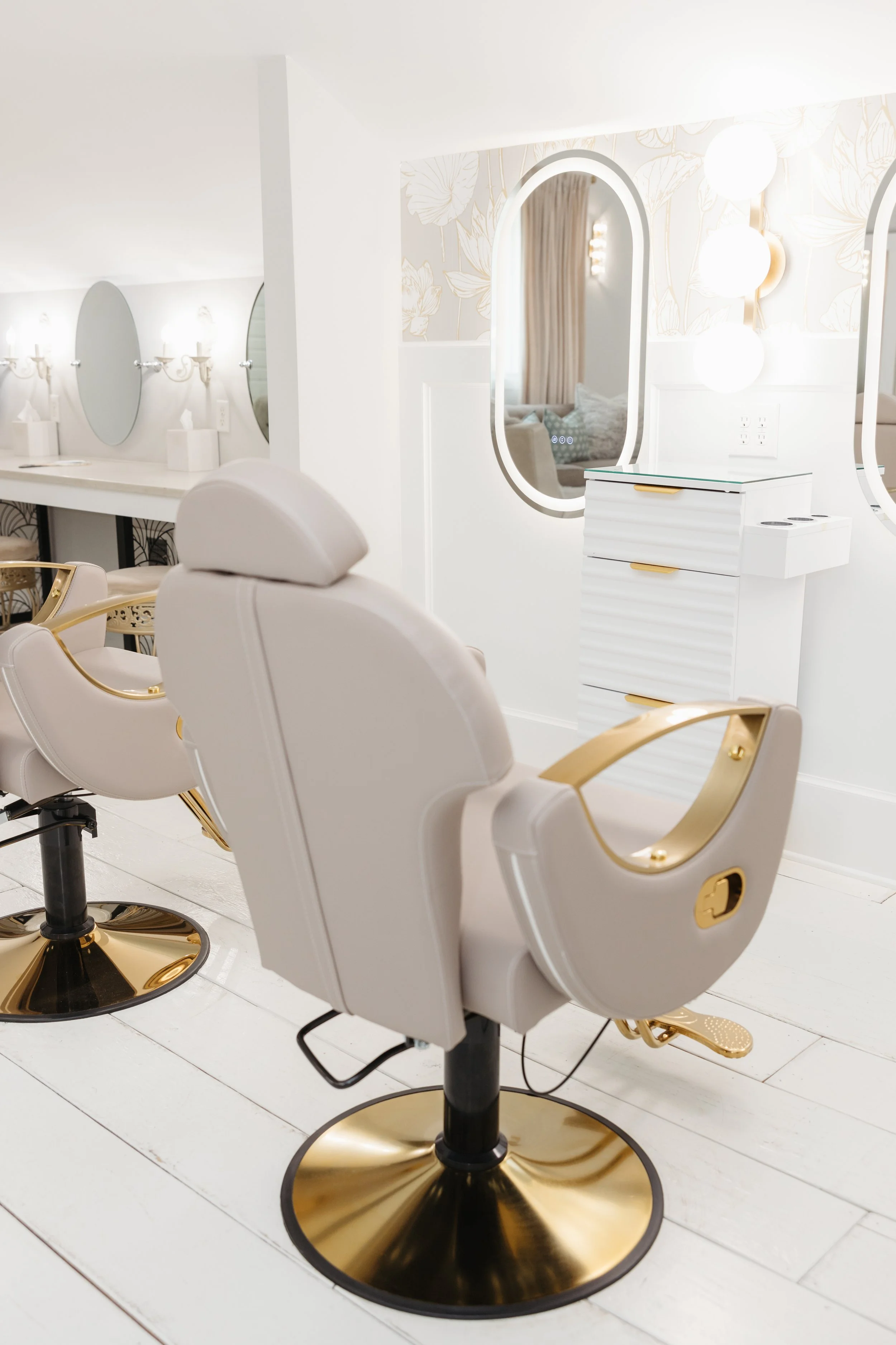 White salon chairs with gold accents arranged in a row in front of mirrors, with a white counter and wall lighting fixtures in the background, perfect for getting ready for your wedding day at Ramble Creek.