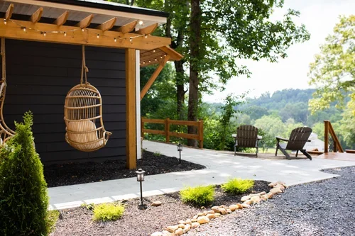 Backyard with a swing chair hanging from a wooden pergola, outdoor seating with two chairs, a table, and a view of trees and hills in the distance.