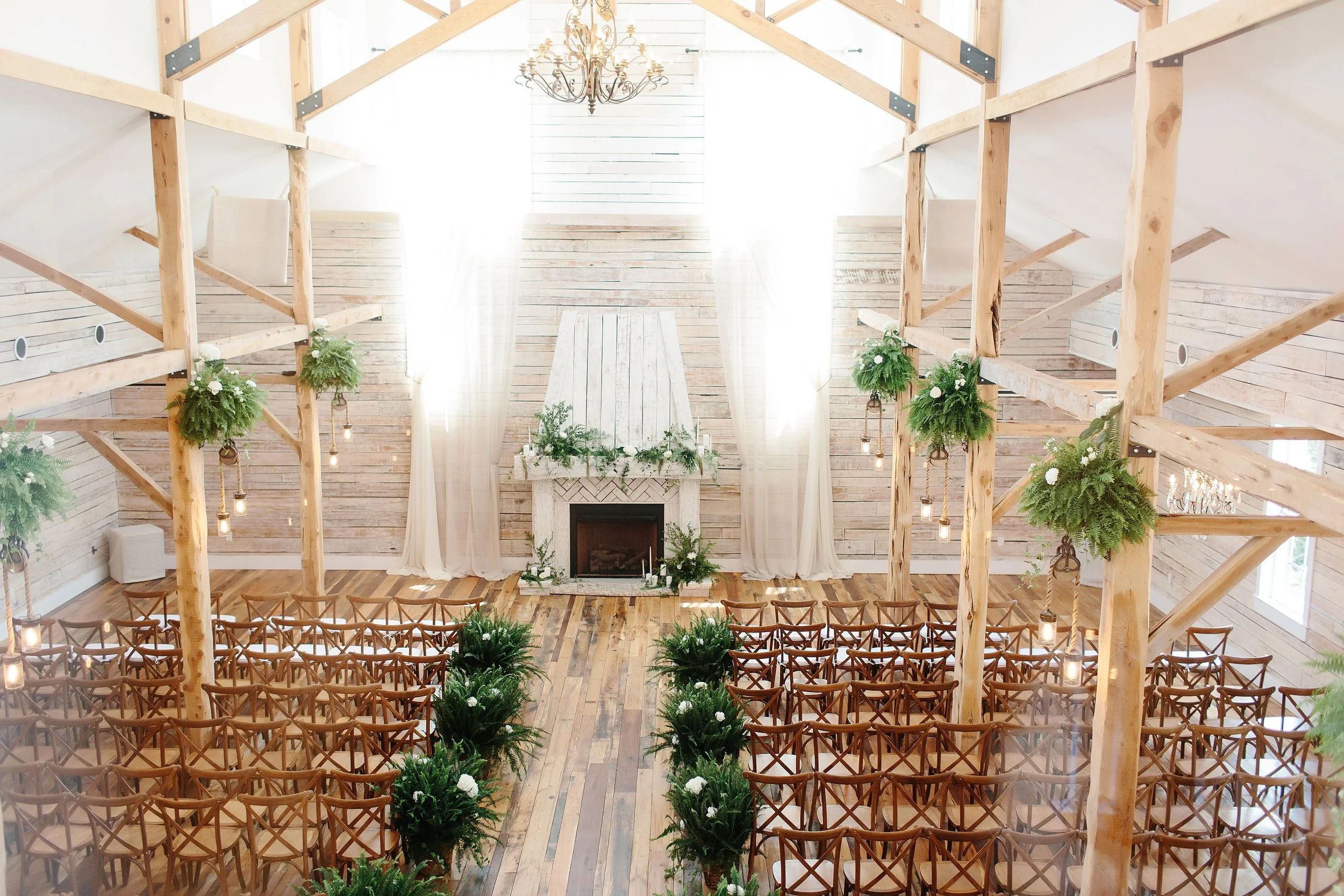 Circular photo of The Grand Hall at Ramble Creek featuring a bright whitewashed barn interior with wooden beams, a central aisle lined with lush greenery, rows of wooden chairs, and a fireplace altar beneath a chandelier