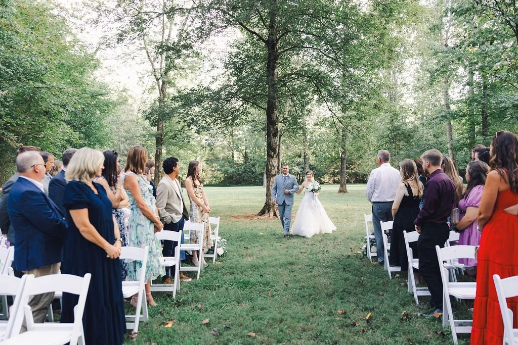 A bride and groom walking down the aisle at an outdoor wedding ceremony in a wooded area of Ramble Creek wedding venue with guests standing on either side.