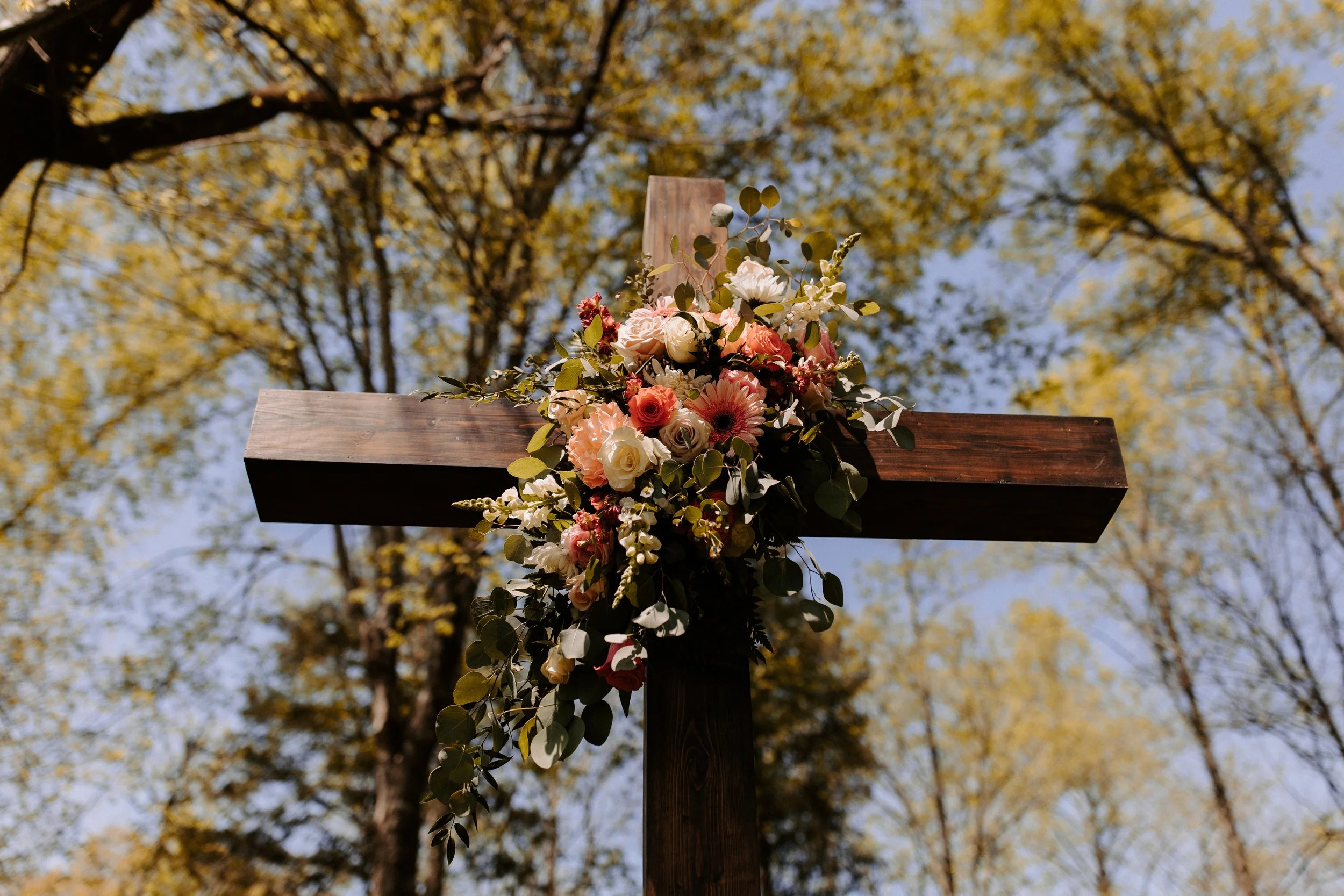 Outdoor ceremony cross adorned with cascading floral arrangements featuring blush roses, white blooms, pink gerbera daisies, snapdragons, eucalyptus, and trailing greenery at Ramble Creek.