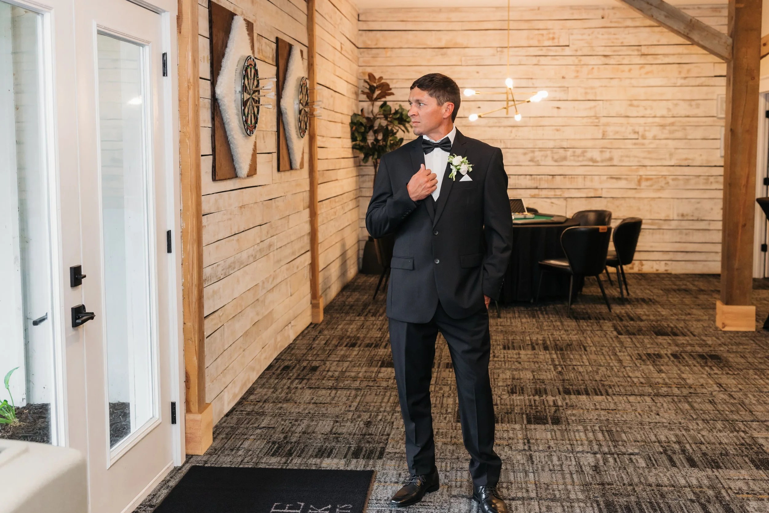 Groom in a black tuxedo with a bow tie and a boutonniere is standing indoors near a door and looking to the side. The room has wood-paneled walls and a patterned carpet.