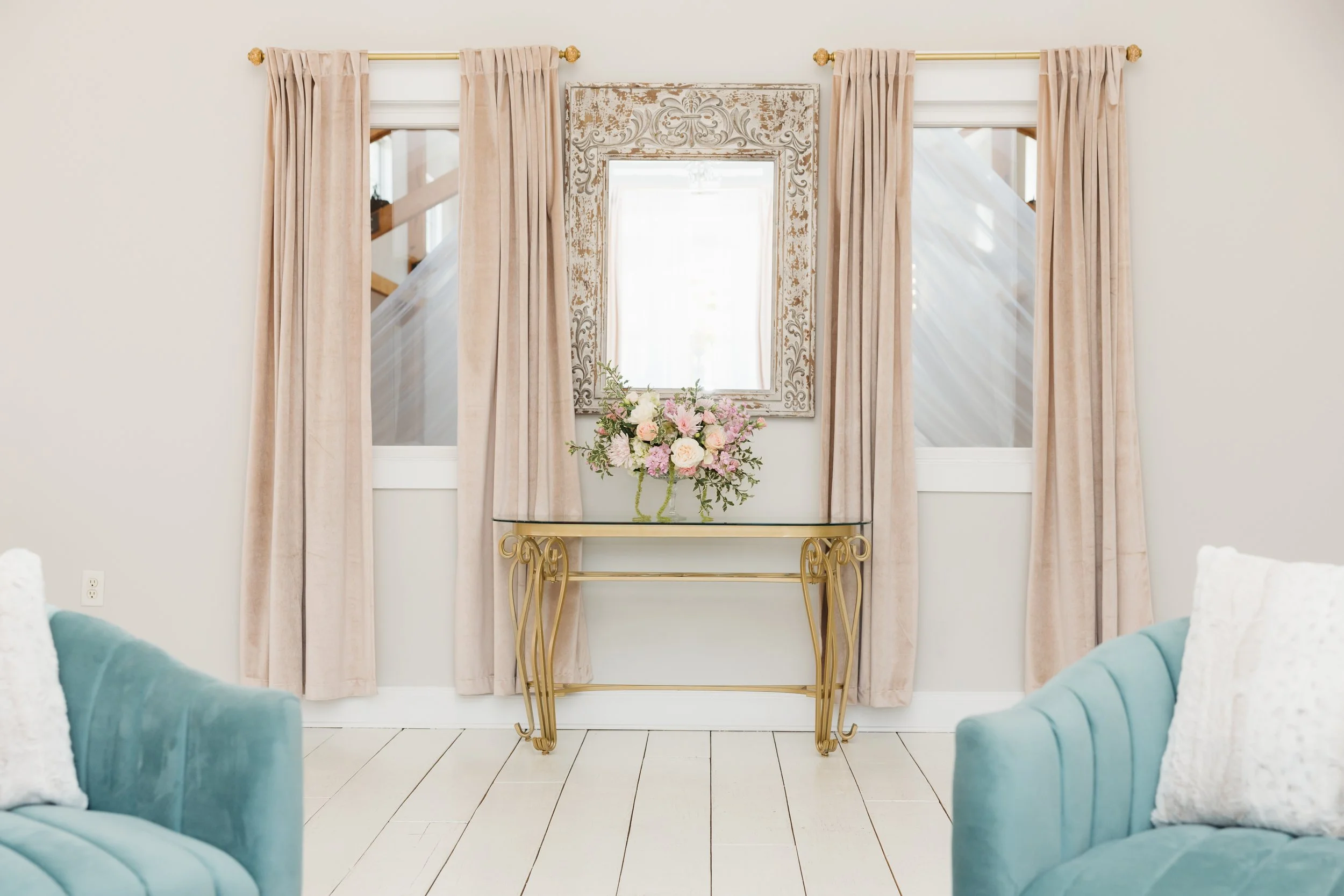 A light-colored room with two teal armchairs on either side of a glass table, decorated with a floral arrangement. Behind the table, there's a distressed, ornate mirror framed with a vintage design. Two windows with beige curtains flank the mirror, a