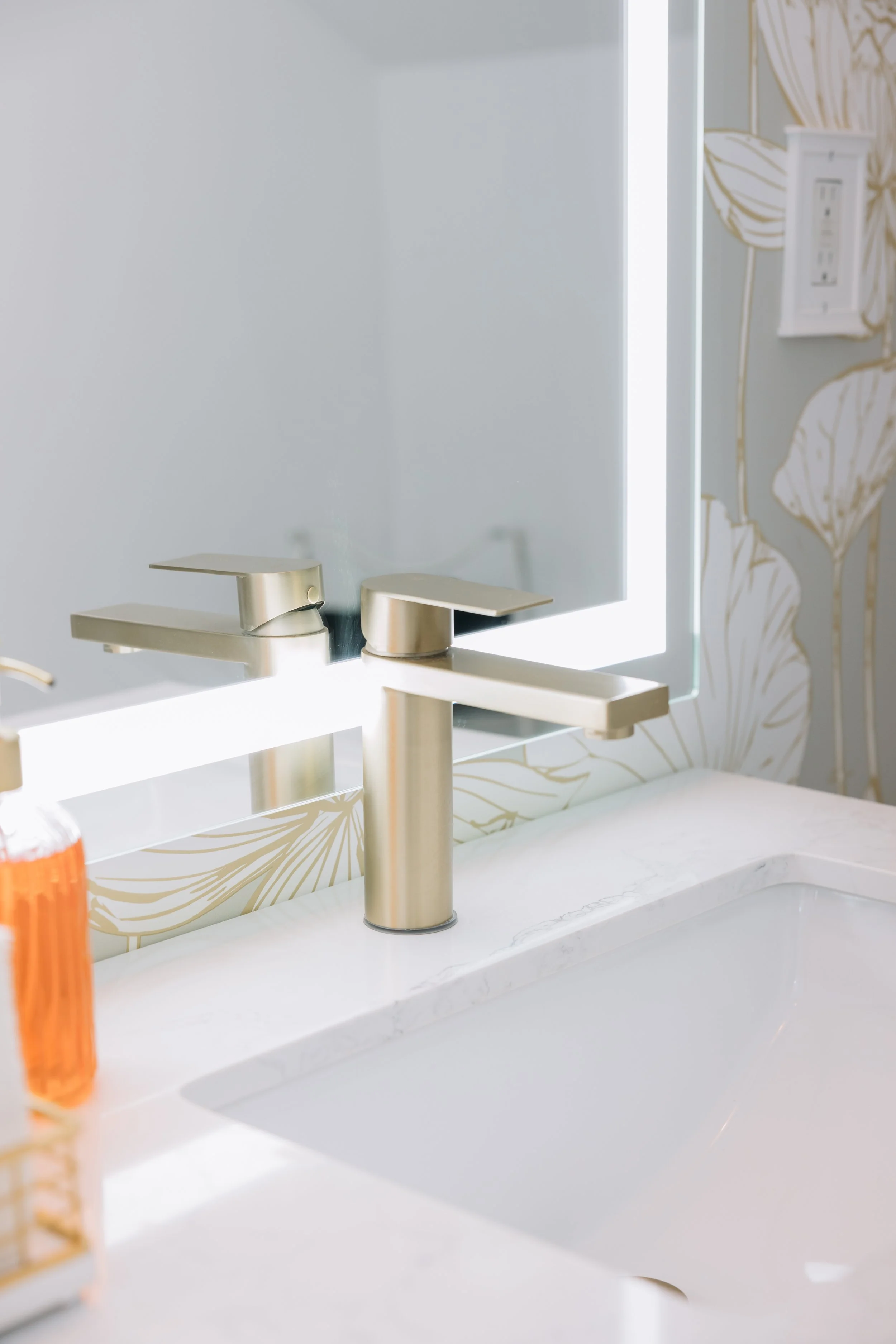 Close-up of bridal suite bathroom sink with a sleek gold faucet, a soap dispenser, and a large mirror reflecting part of the shower or wall. The countertop is white stone, and there is decorative wallpaper with a gold-leaf pattern.