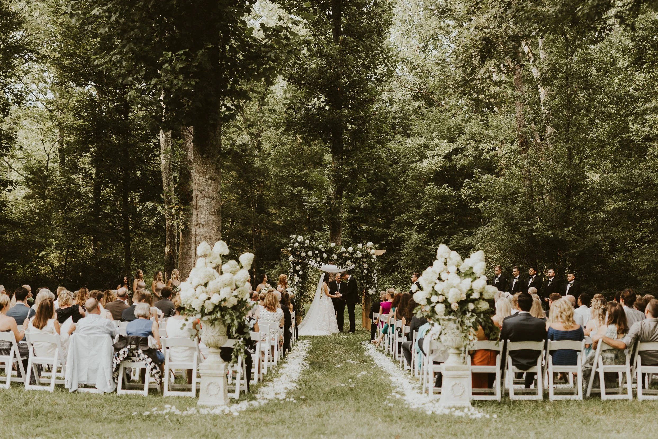 A wedding ceremony taking place outdoors in a forested area with the bride and groom exchanging vows under an arch decorated with flowers, surrounded by seated guests and floral arrangements. Located in east TN at all inclusive wedding venue.