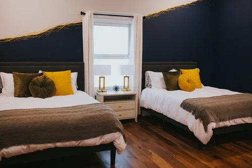 Two beds in a bedroom with dark blue walls and a white window in the middle, decorated with yellow and brown pillows and brown blankets, and a white nightstand between them.