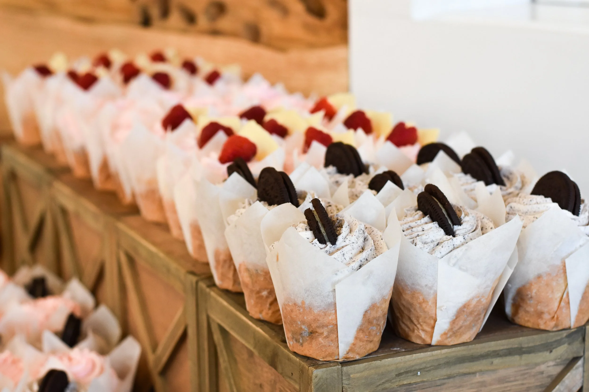 Display of various wedding cupcakes with different toppings, arranged on a wooden shelf at Ramble Creek wedding reception.