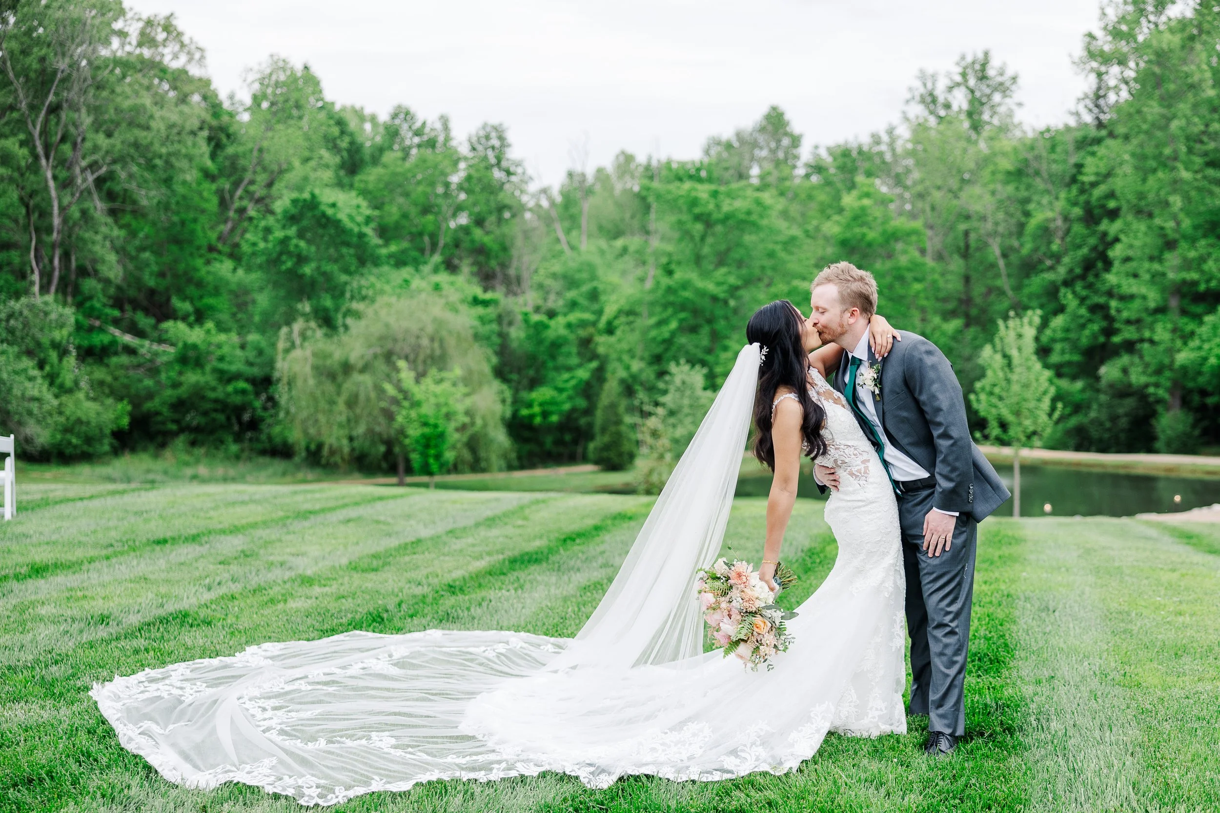 A newlywed couple kissing outdoors on green grass with trees in the background. The bride is wearing a white lace wedding dress with a long veil and holding a bouquet of flowers, while the groom is dressed in a dark suit.