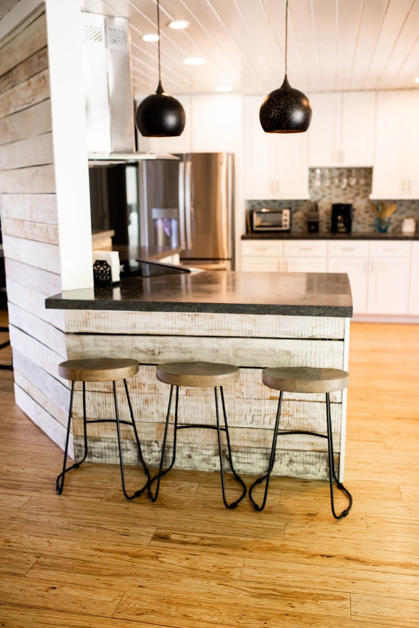 Kitchen island at the Manor onsite accommodation at Ramble Creek, featuring rustic wood paneling, black pendant lights, bar stools, white cabinetry, and warm wood floors.