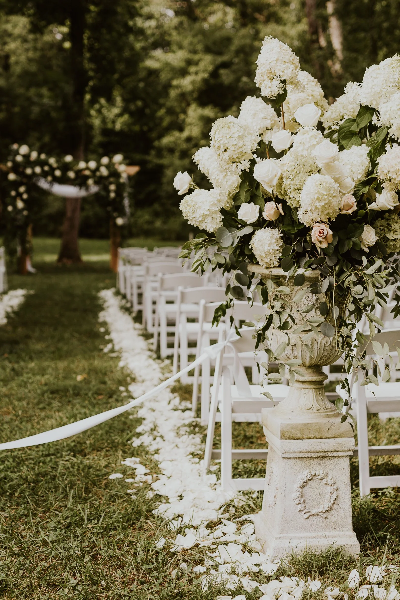 Elegant outdoor wedding setup with white chairs along a grassy aisle, decorated with a large floral arrangement of white hydrangeas and roses on a stone pedestal in a lush green garden at Ramble Creek all-inclusive wedding venue.