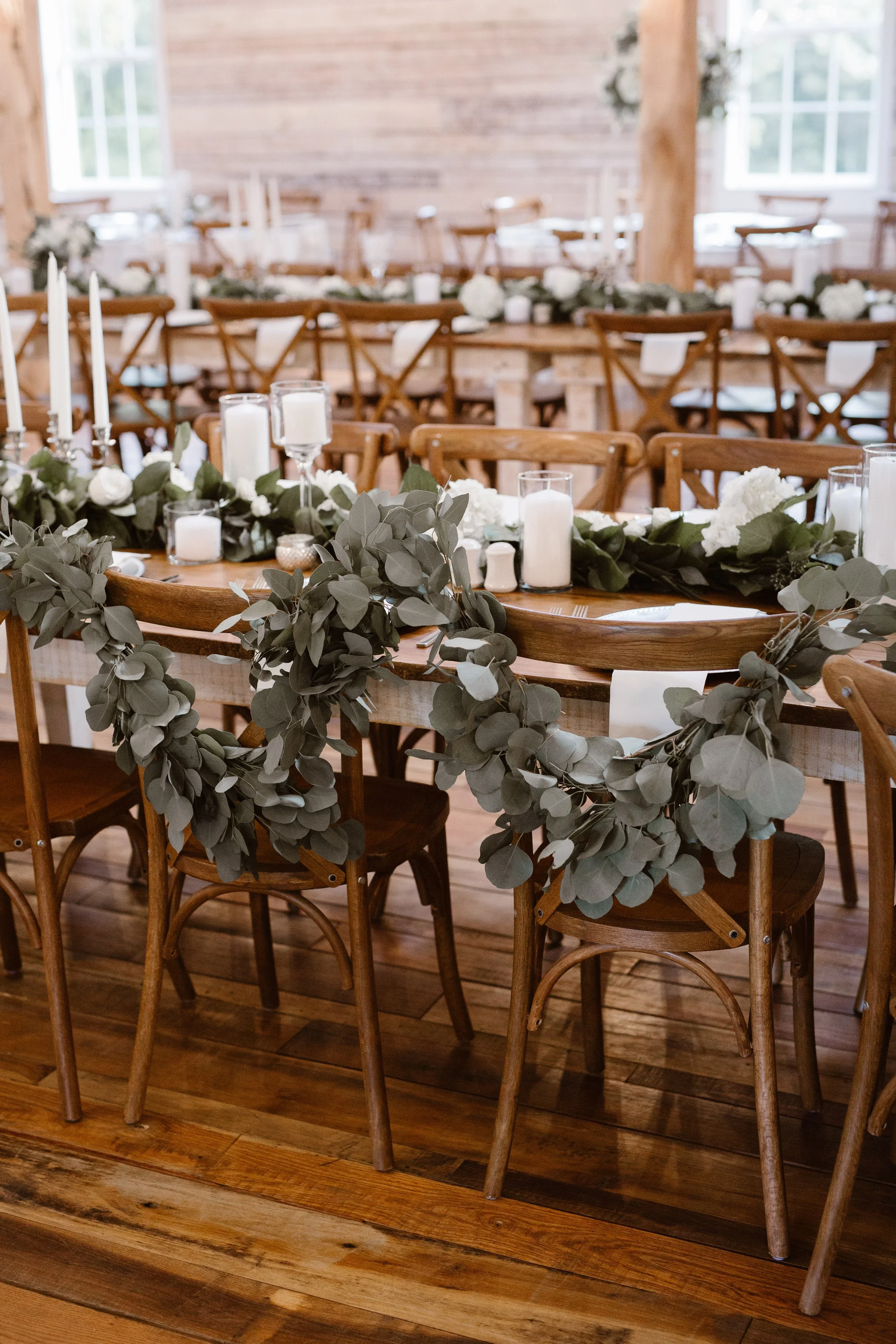 Ramble Creek wedding reception with wood cross-back chairs, greenery garlands, white floral accents, and candlelit tables in an elegant indoor setting.