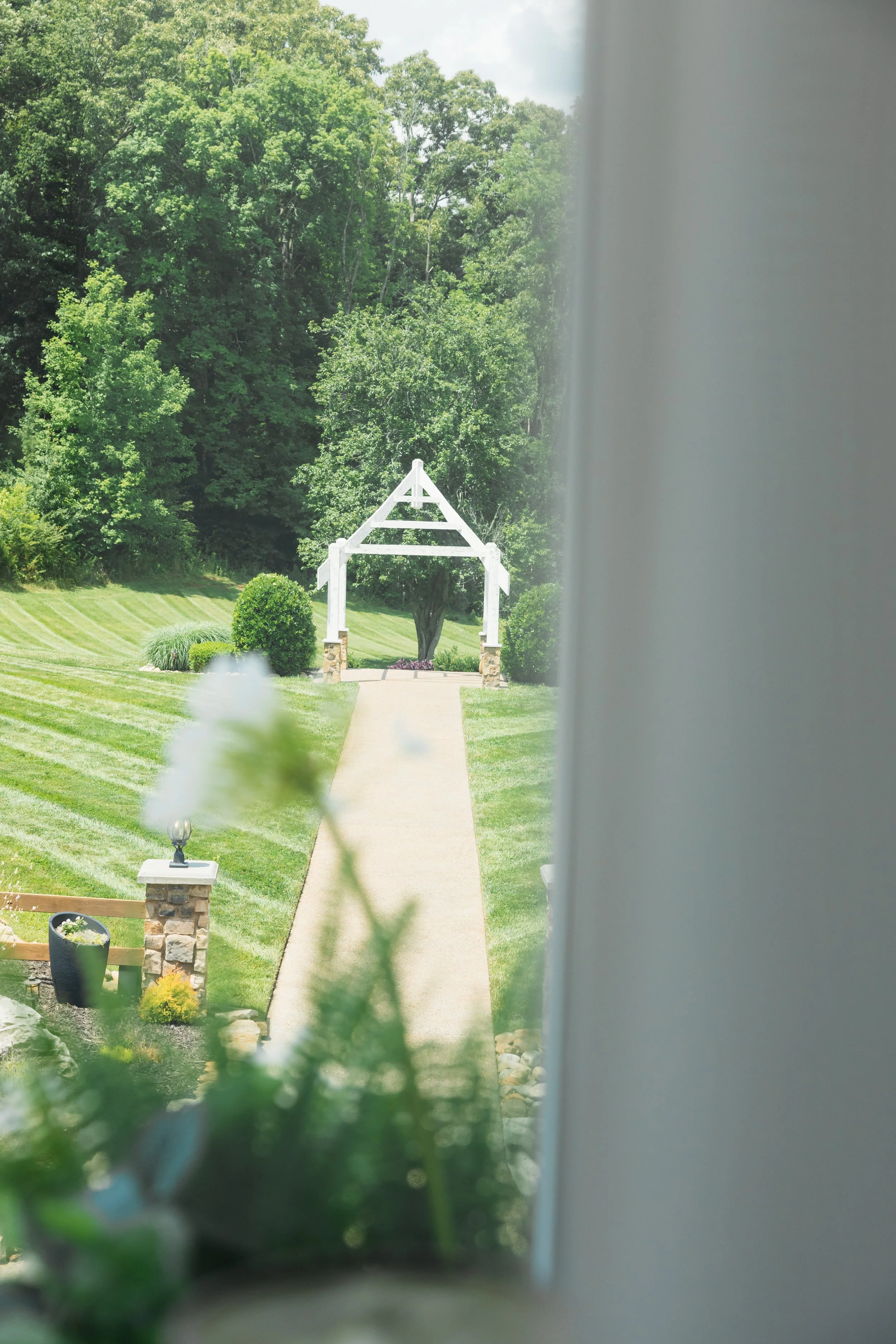 A view of a well-maintained outdoor ceremony site lawn and a white wooden archway leading to a wooded area, with a sidewalk and decorative landscape elements in the foreground, partially obscured by a wall or curtain.