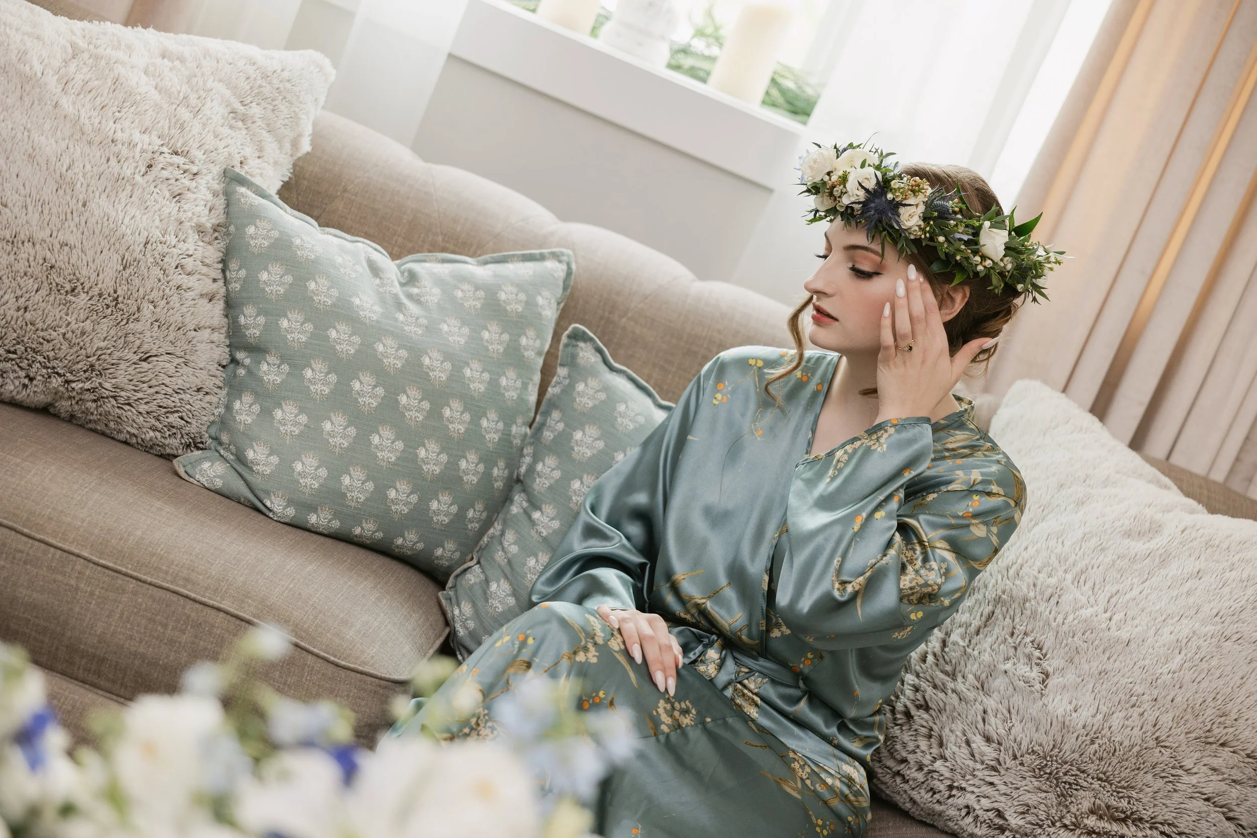 A bride wearing a floral crown and a silk robe sitting on a beige couch in a bright room with large windows, surrounded by soft pillows and a furry blanket, as she gets ready for her wedding day at Ramble Creek wedding venue.