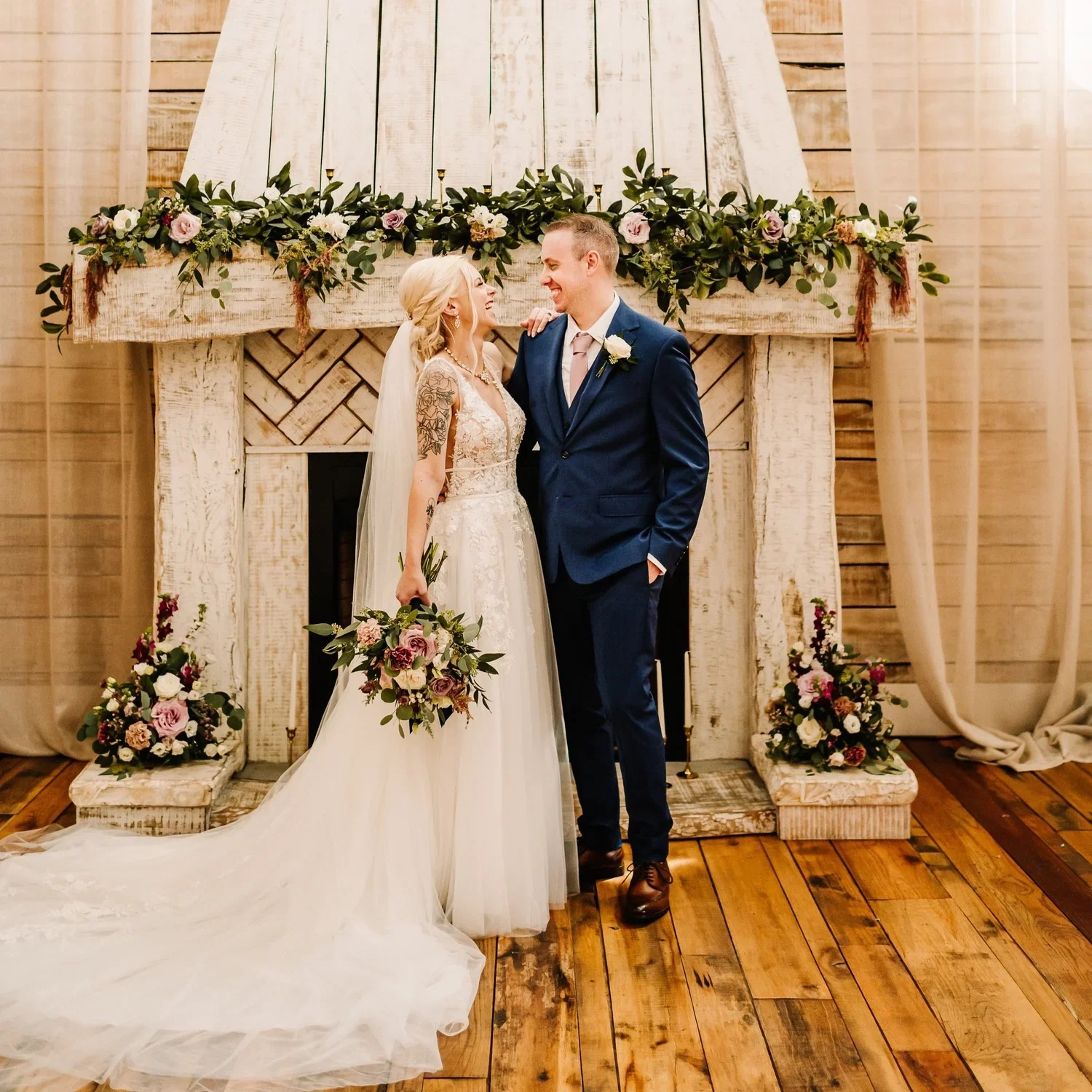 A bride and groom standing in front of a rustic fireplace decorated with flowers during a wedding ceremony. The bride has blonde hair, tattoos on her arm, and is holding a bouquet. The groom has short hair and is wearing a navy suit with a white shirt and a boutonniere.