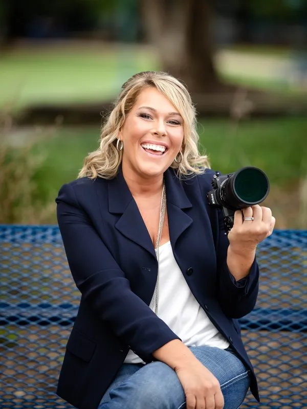 A woman with blonde, wavy hair, wearing a navy blazer, white top, and jeans, sitting on a bench outdoors, smiling and holding a camera.