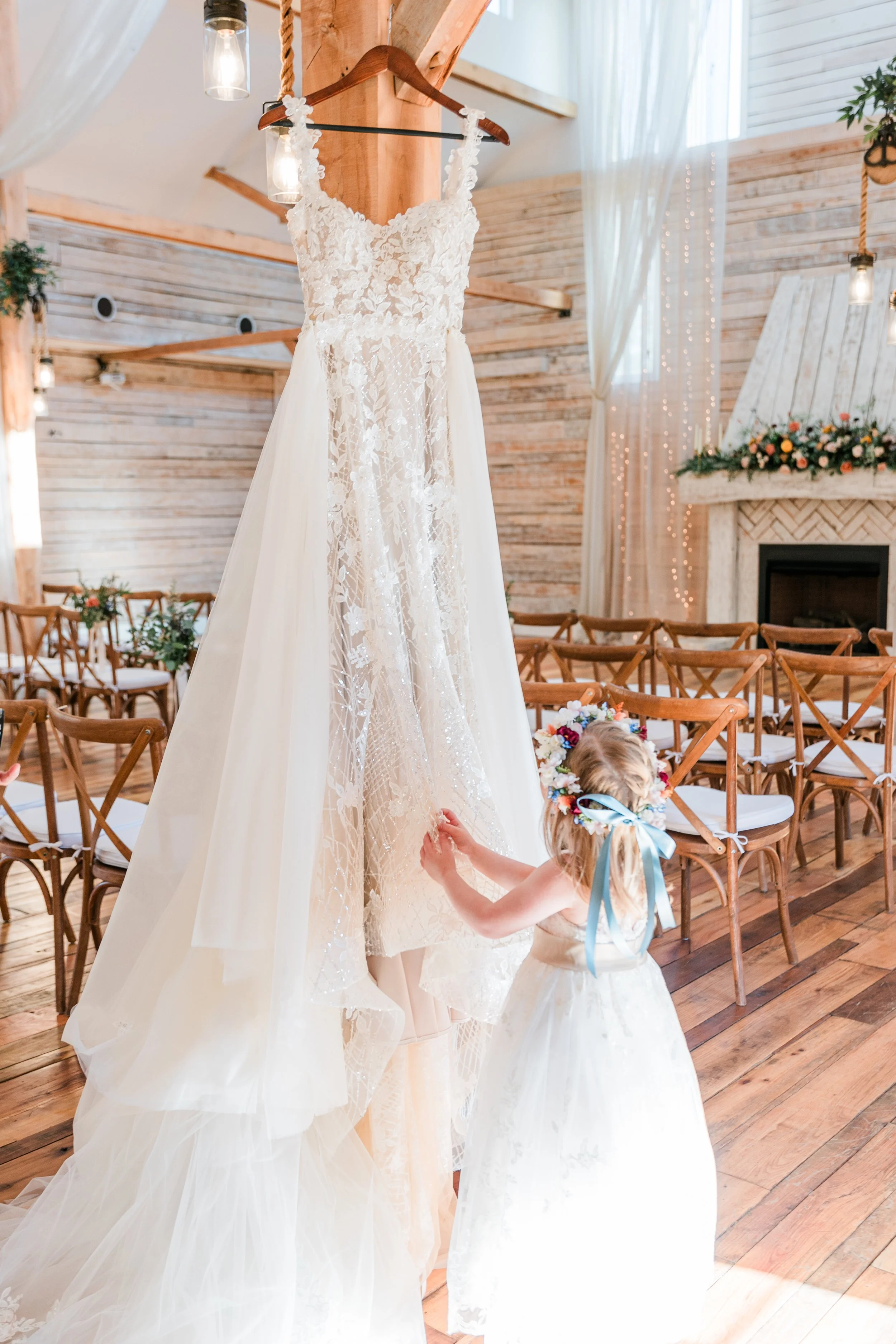 A young girl wearing a flower crown and a light-colored dress is adjusting a white lace wedding dress hanging from a wooden beam in a beautiful TN wedding venue with wooden chairs, a fireplace, floral arrangements, and string lights.