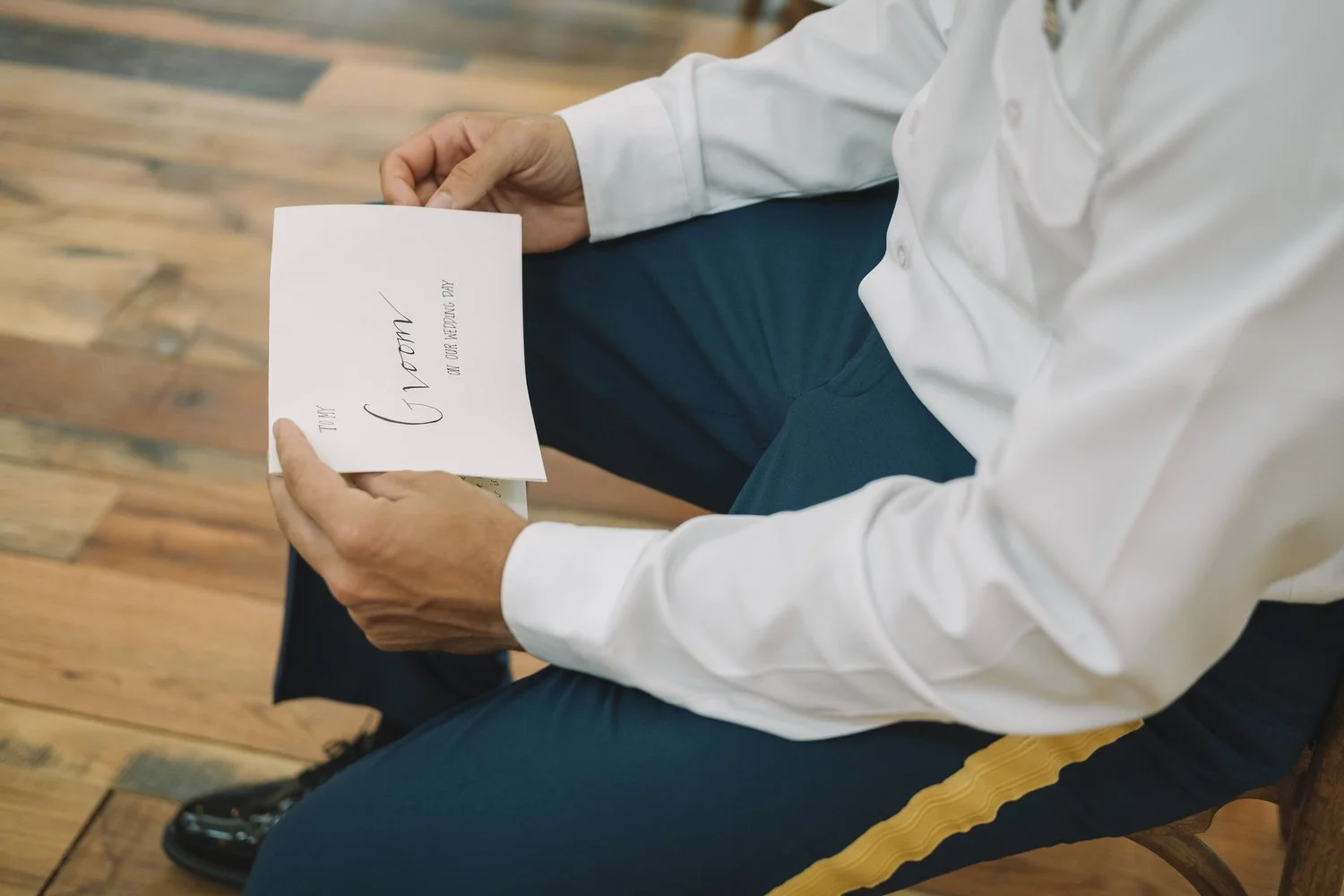 Groom sitting on a wooden chair in the Grand Hall, holding a wedding note from his soon-to-be wife.