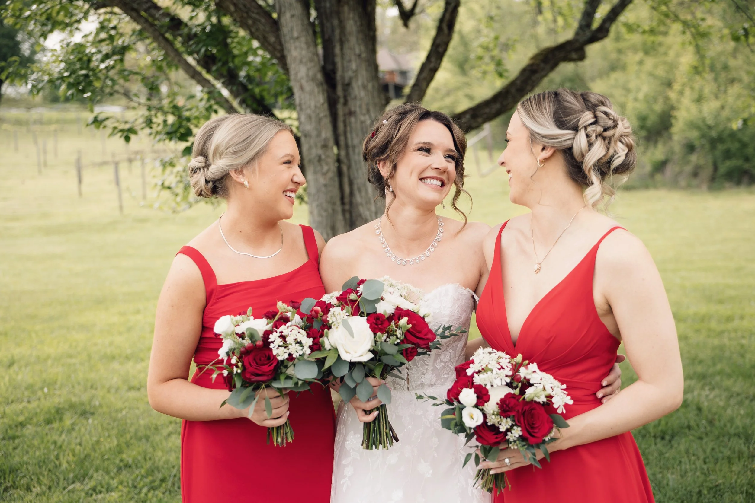 Two women in red dresses and a bride in a white wedding gown standing outdoors under a tree, smiling and holding bouquets of flowers at a wedding in Tennessee.
