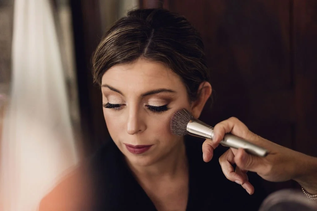 Makeup artist applying blush to a bride with soft glam wedding makeup and a sleek low hairstyle