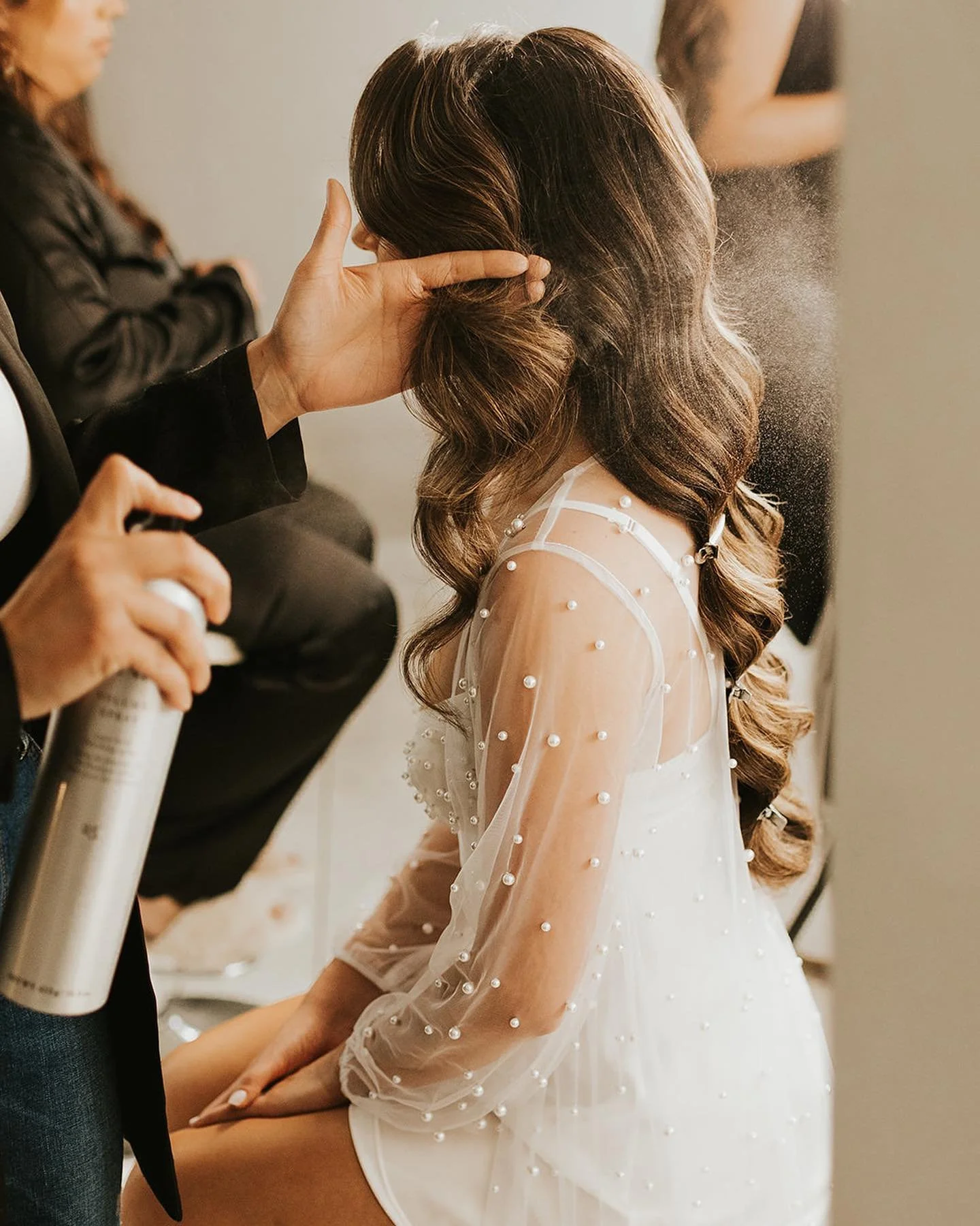 A bride with long, wavy brown hair in a white dress with pearl embellishments is getting her hair styled. A Ramble Creek in-house stylist is holding a spray bottle and a hairbrush, working on her hair in the bridal suite.
