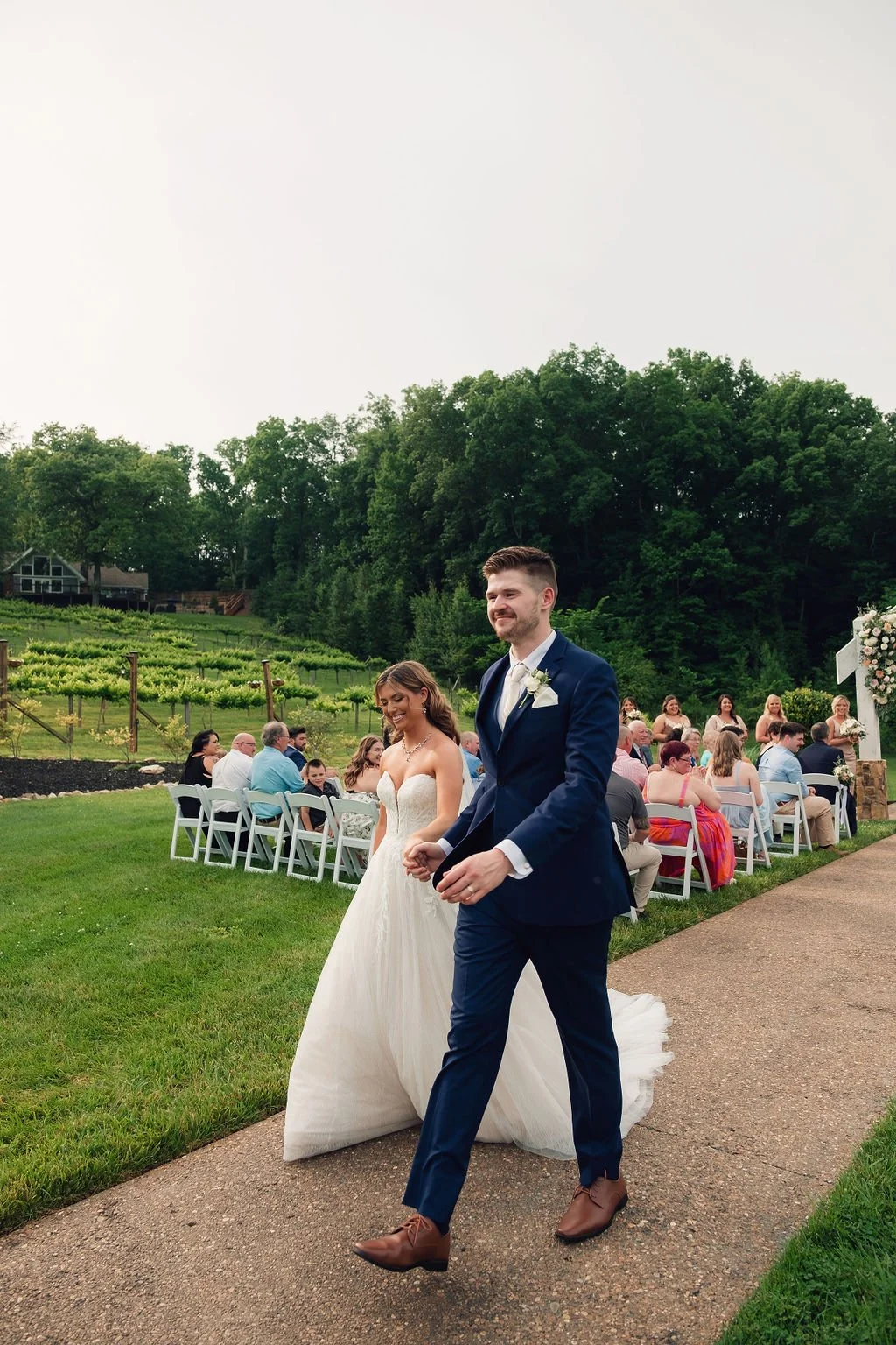 A newlywed couple walking hand in hand outdoors during their outdoor wedding ceremony in TN mountains, with guests seated on white chairs behind them on a grassy area, surrounded by trees and a scenic landscape.