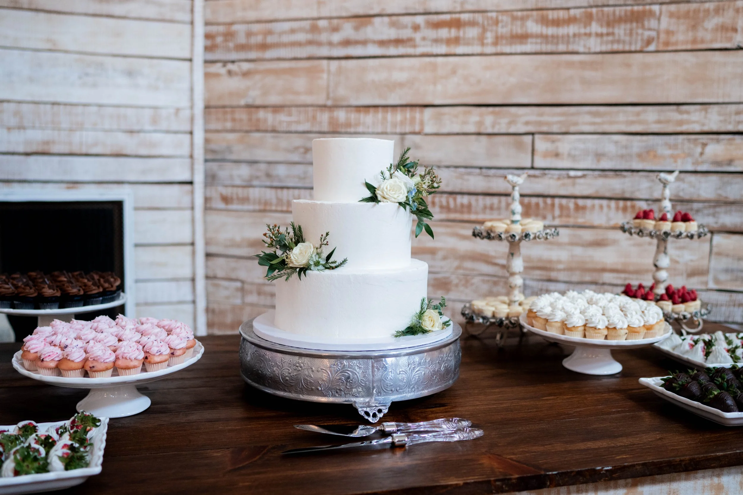 A three-tier white wedding cake decorated with white roses and greenery, surrounded by various cupcakes and desserts on display tables with a wooden wall background.