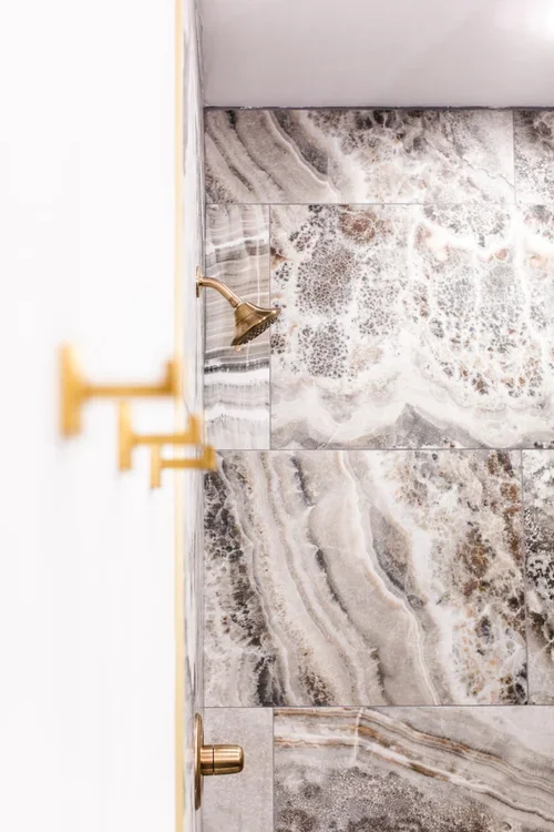 View of a shower with marble tiles, a gold showerhead, and a white door partially open.
