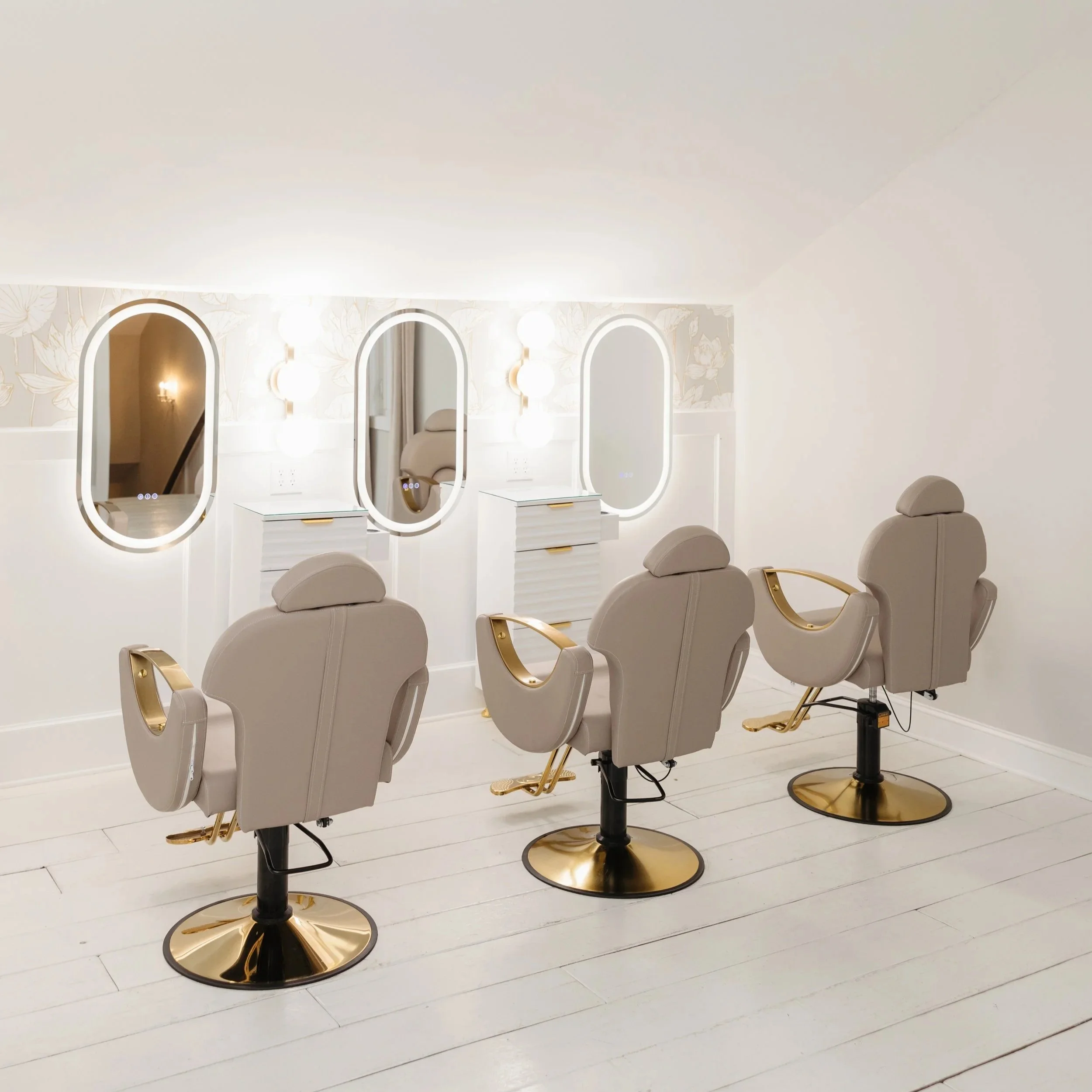 In Ramble Creek bridal suite, a row of three oval mirrors with white frames and vanity tables beneath, illuminated by round wall-mounted lights, and beige salon chairs with gold accents in front of each mirror.