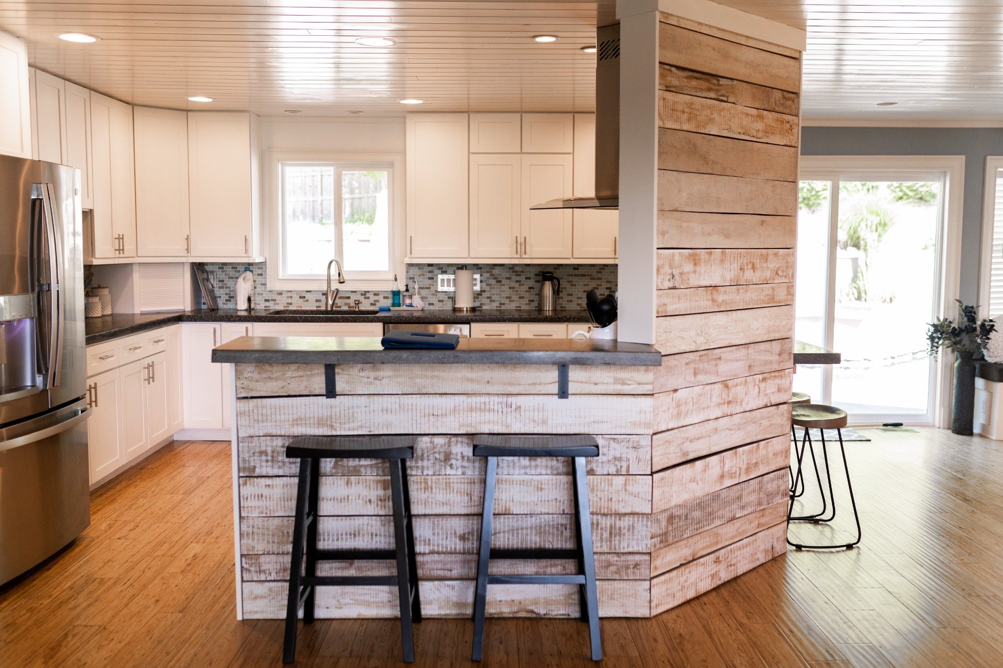 Kitchen at the Manor onsite accommodation at Ramble Creek, featuring white cabinetry, a rustic wood breakfast bar, black stools, stainless steel appliances, and warm wood floors.