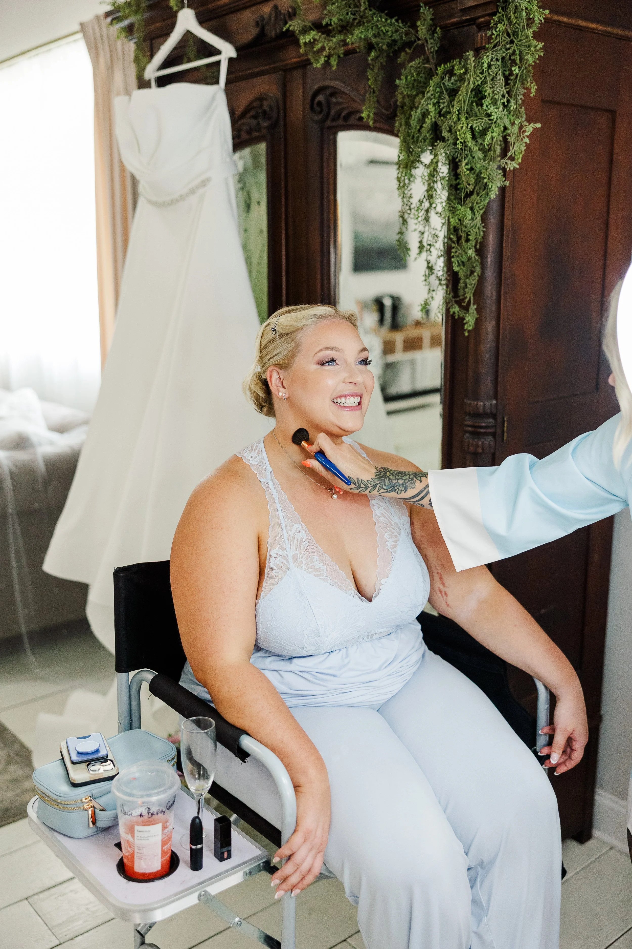 Smiling bride getting blush applied with her wedding dress hanging behind her