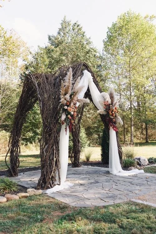 Circular photo of the Vine Arbor ceremony site at Ramble Creek featuring a rustic outdoor arbor made of intertwined vines and draped white fabric, accented with floral arrangements and pampas grass, set among tall trees and natural greenery