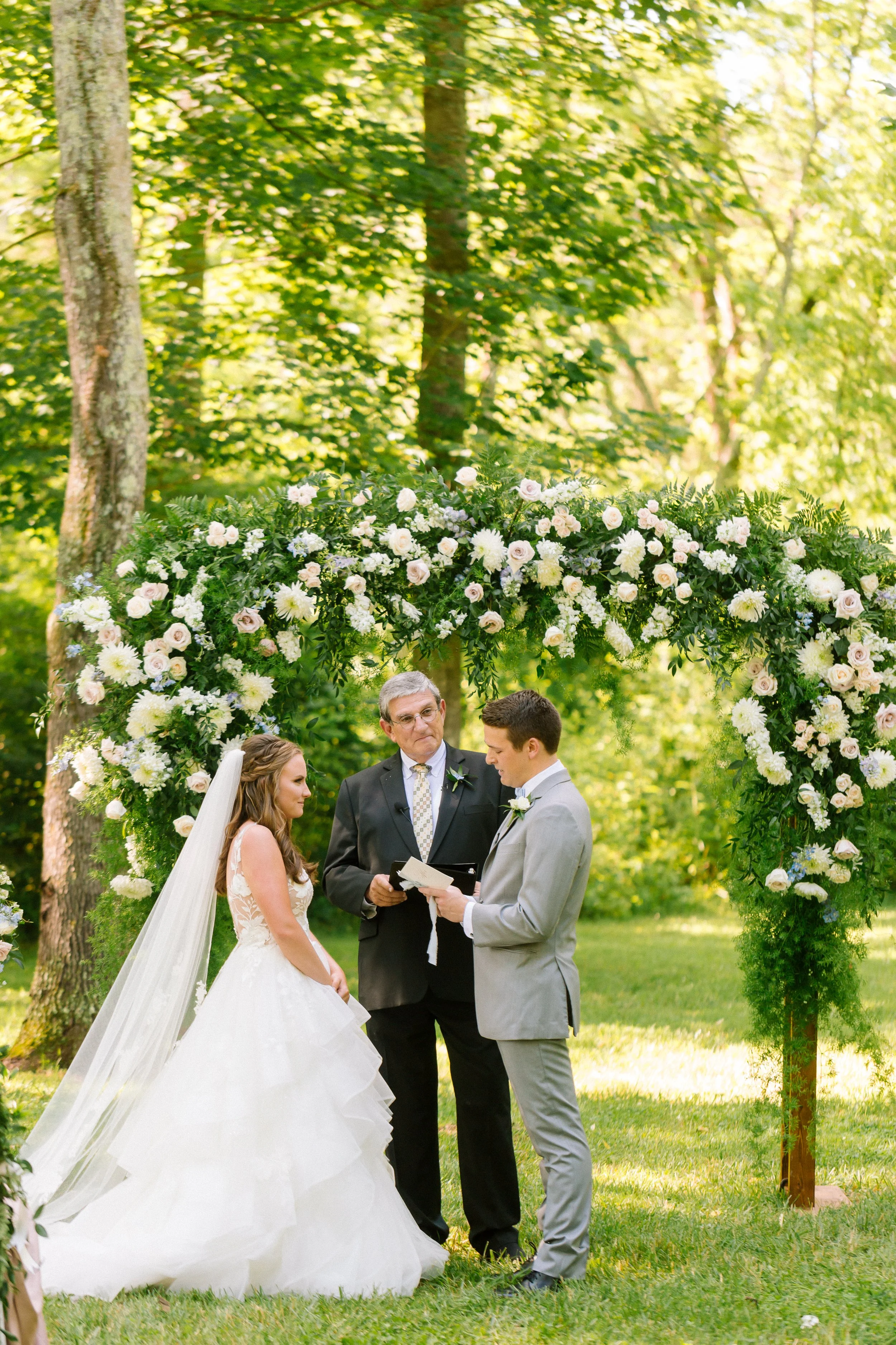 A wedding ceremony outdoors under a floral arch, with a bride and groom facing each other, an officiant holding a book, and a bridesmaid with a long veil. Trees and sunlight in the background.