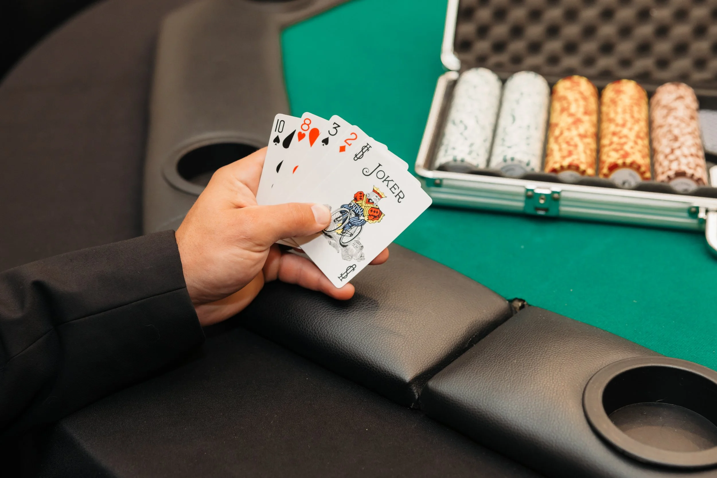 A hand holding five playing cards, including a 10 of spades, 8 of spades, 3 of diamonds, and two jokers, with poker chips and a poker case in the background.