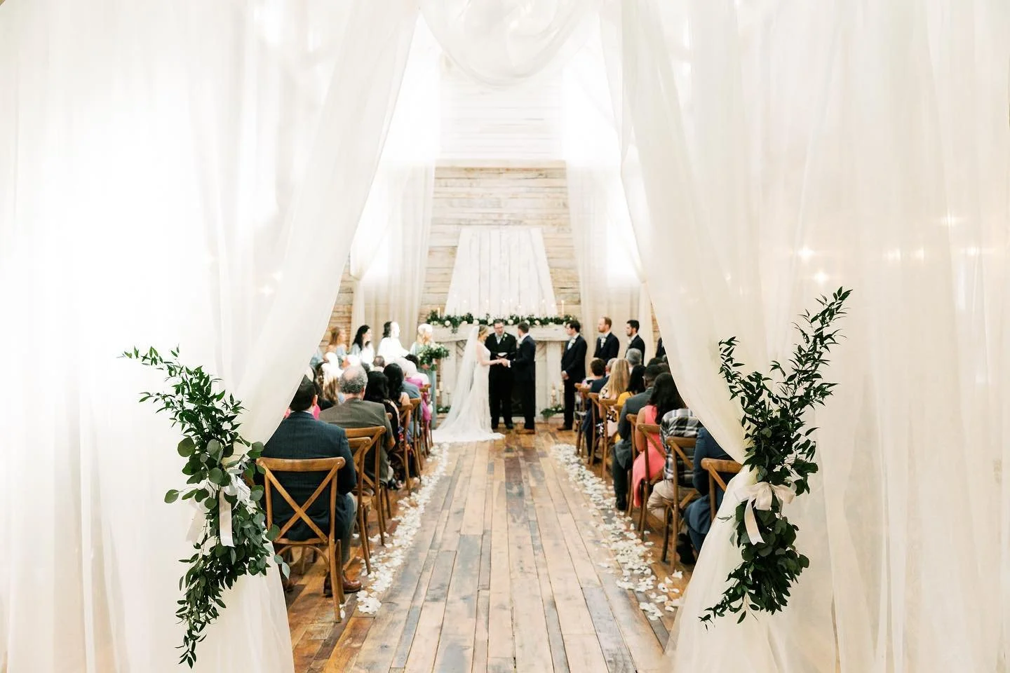 A wedding ceremony inside a rustic wooden venue, viewed through an archway decorated with greenery. The bride and groom are standing at the altar, exchanging vows, with guests seated on either side.
