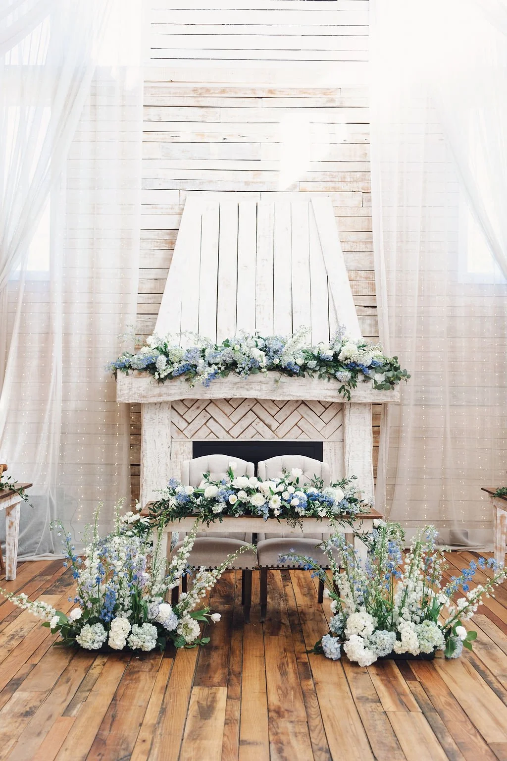 Romantic ceremony floral installation with cascading blue and white blooms surrounding sweetheart chairs and a fireplace mantel at Ramble Creek, styled inside the Grand Hall with soft drapery and rustic wood accents.