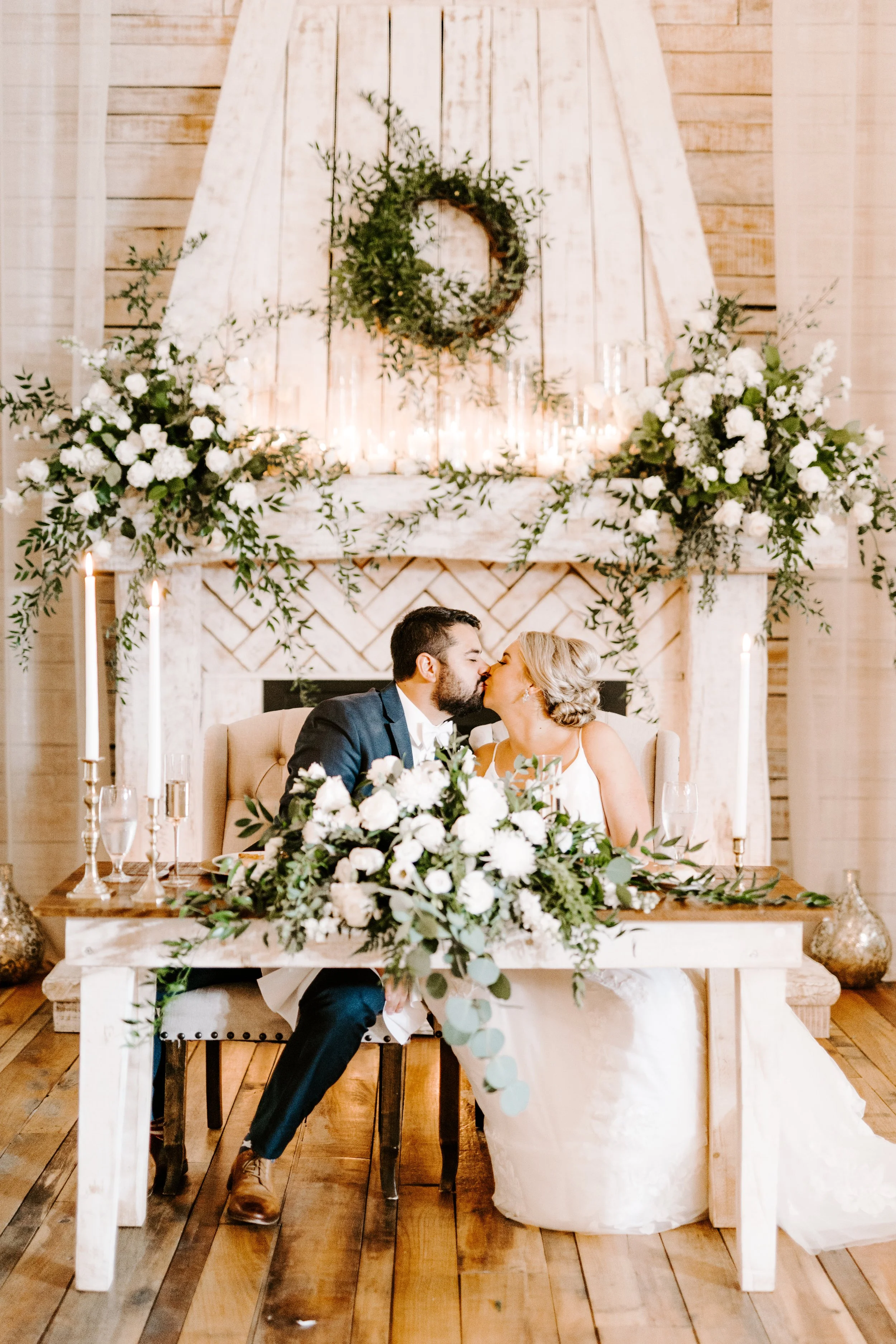 Bride and groom kissing at sweetheart table in The Grand Hall, surrounded by lush white and green floral