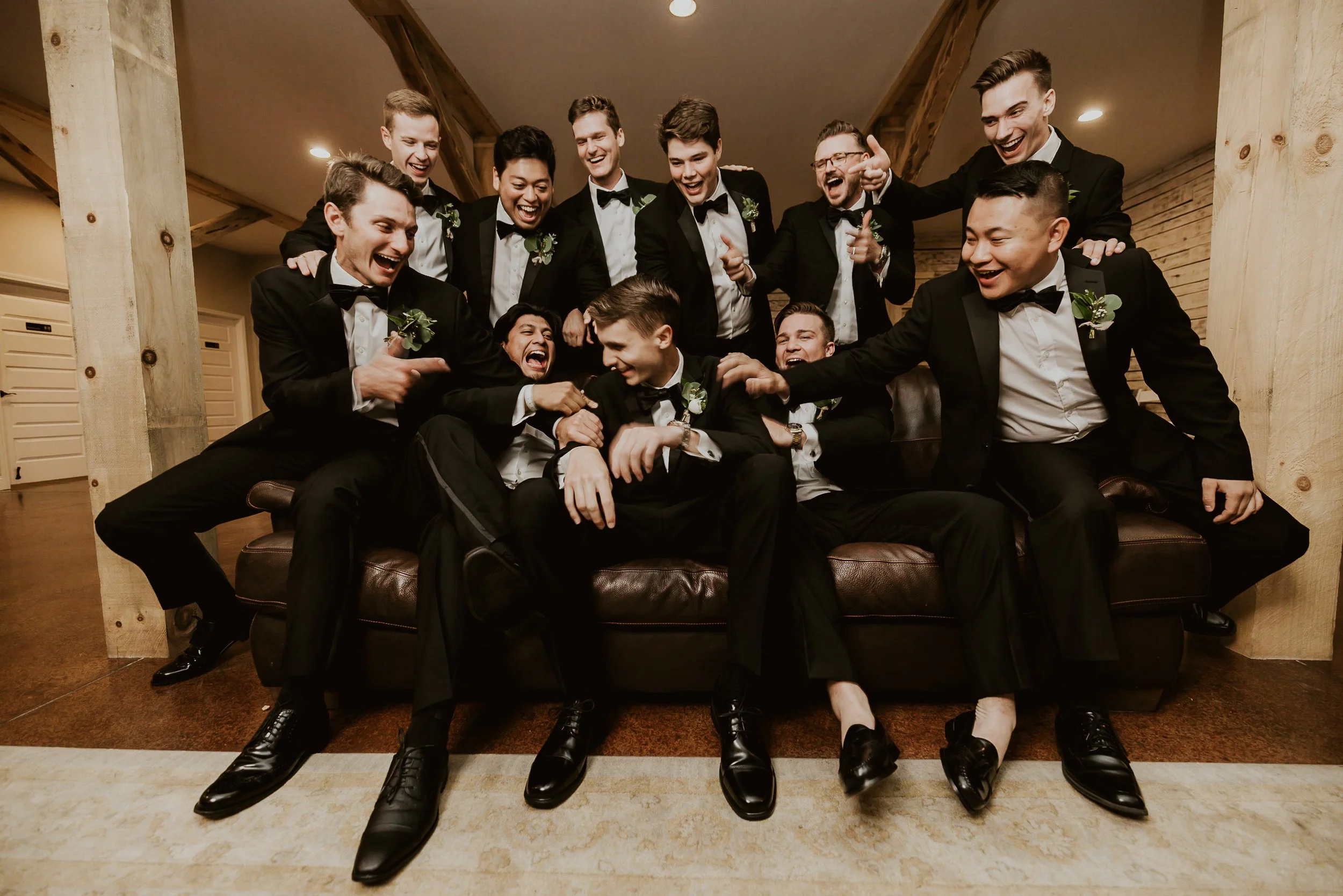 A group of men dressed in tuxedos, sitting and standing, laughing and smiling together in a celebratory setting.