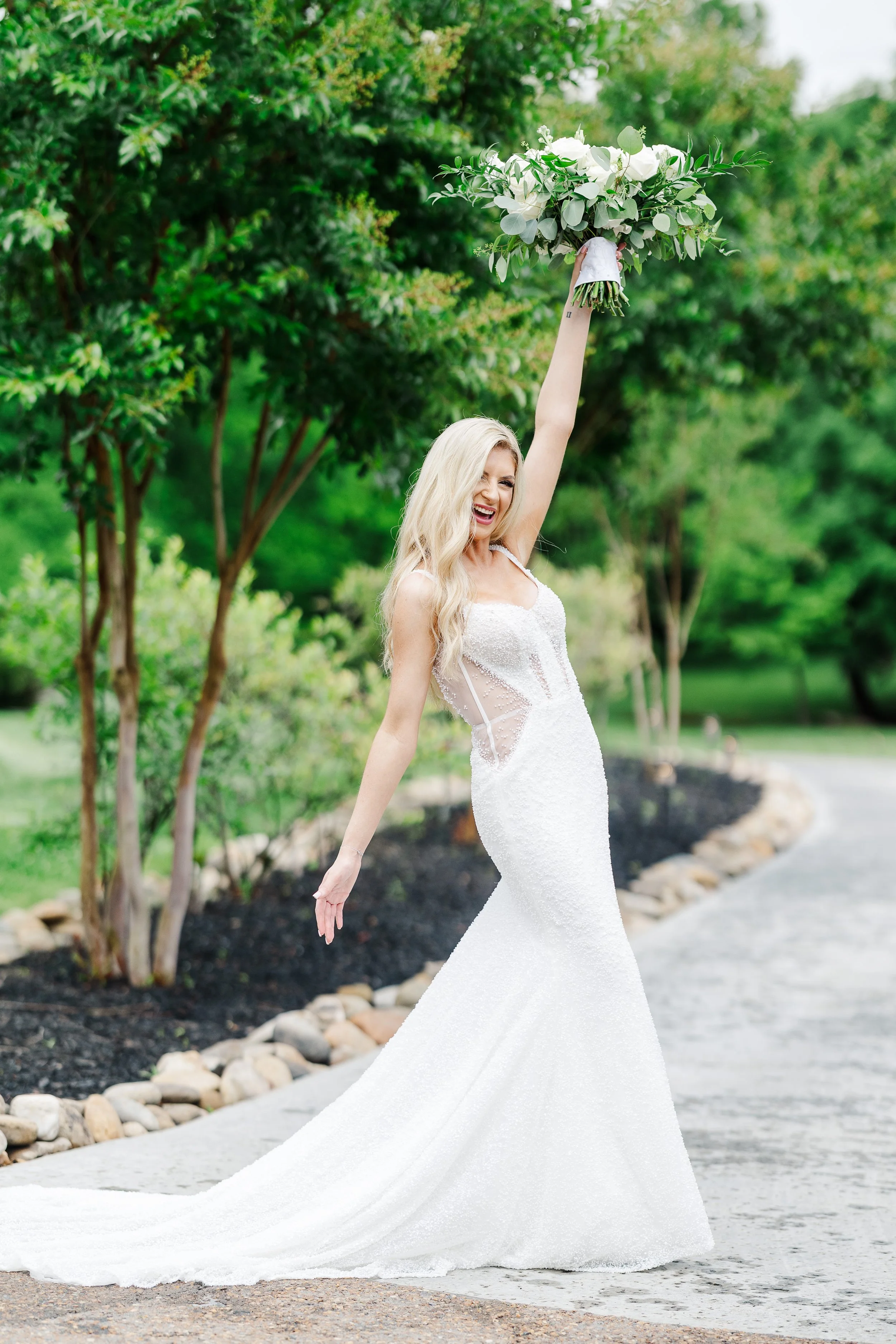 Happy bride in a fitted white wedding dress raising her bouquet in celebration on a garden path at Ramble Creek all-inclusive wedding venue in East TN